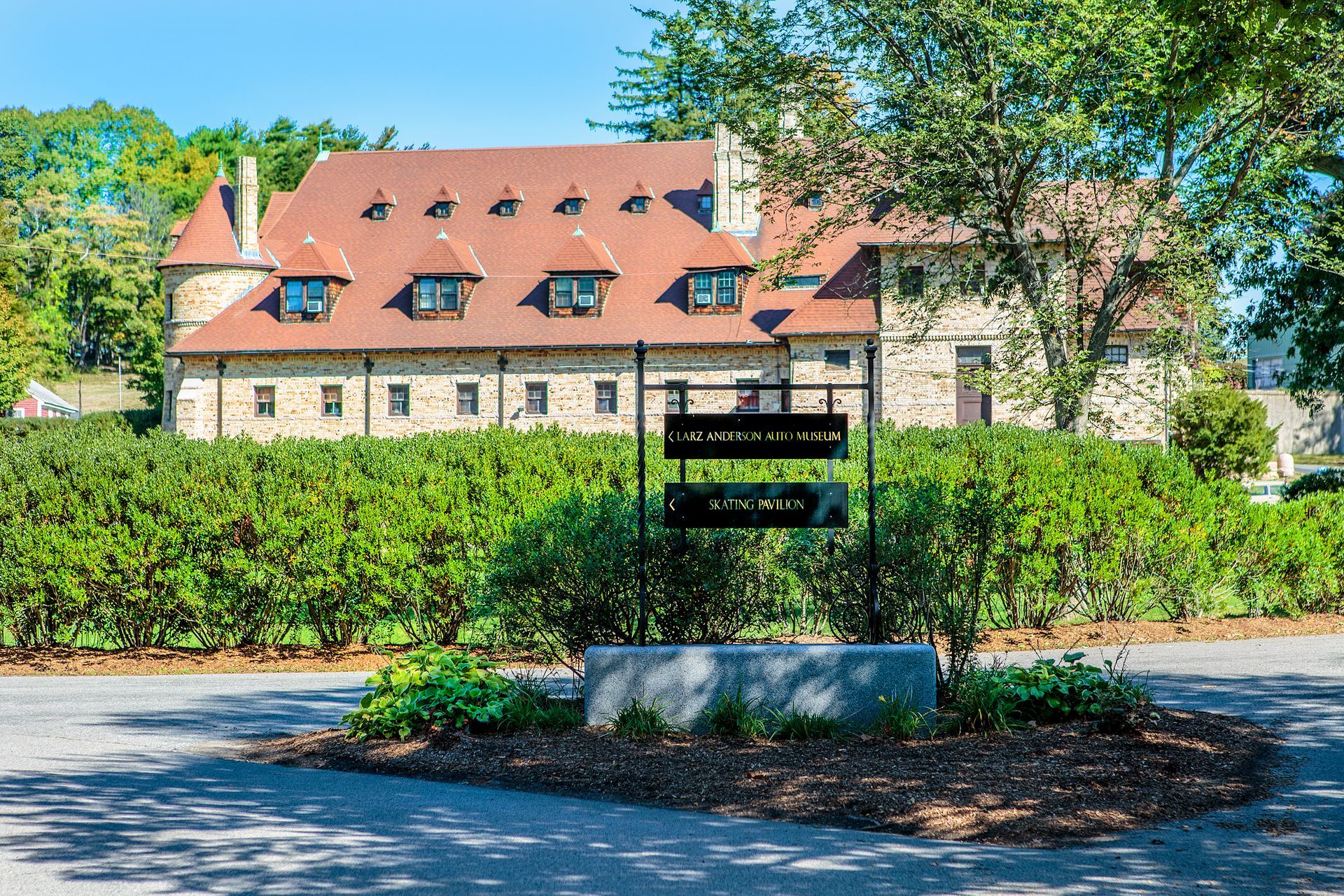 Stone building with red roof, behind a hedge. Sign in front, on gravel.
