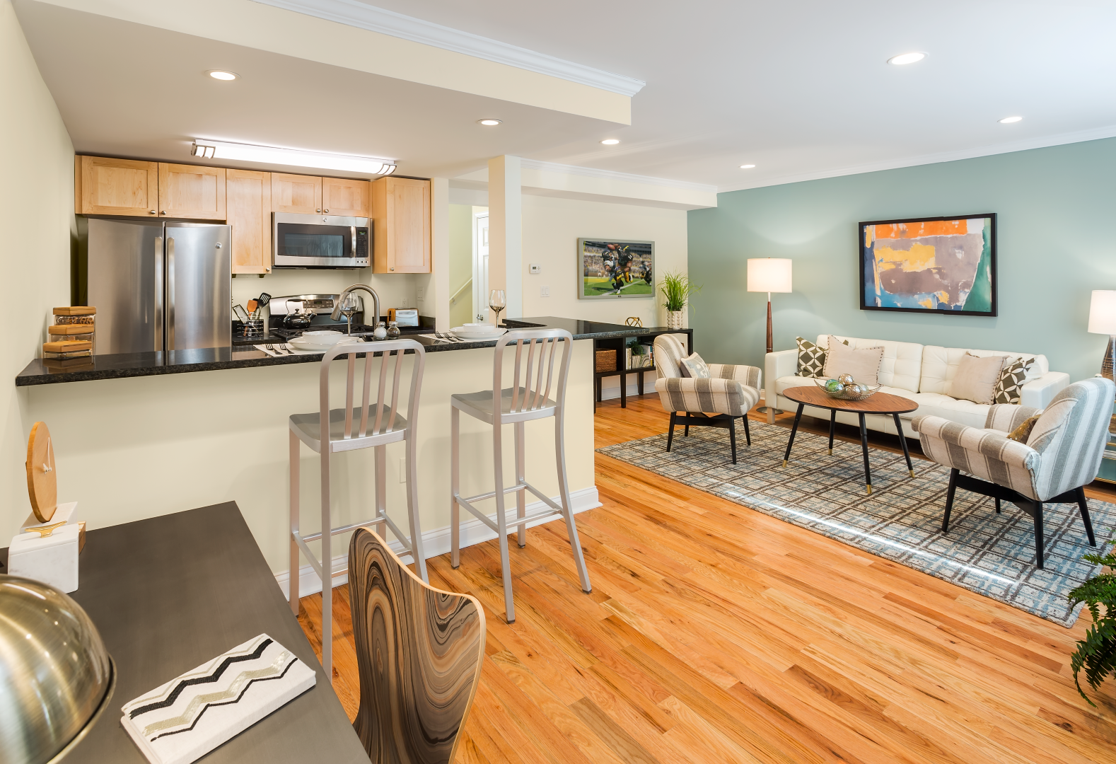 Open-concept living room and kitchen with hardwood floors, white bar stools, and a gray sofa with patterned pillows.