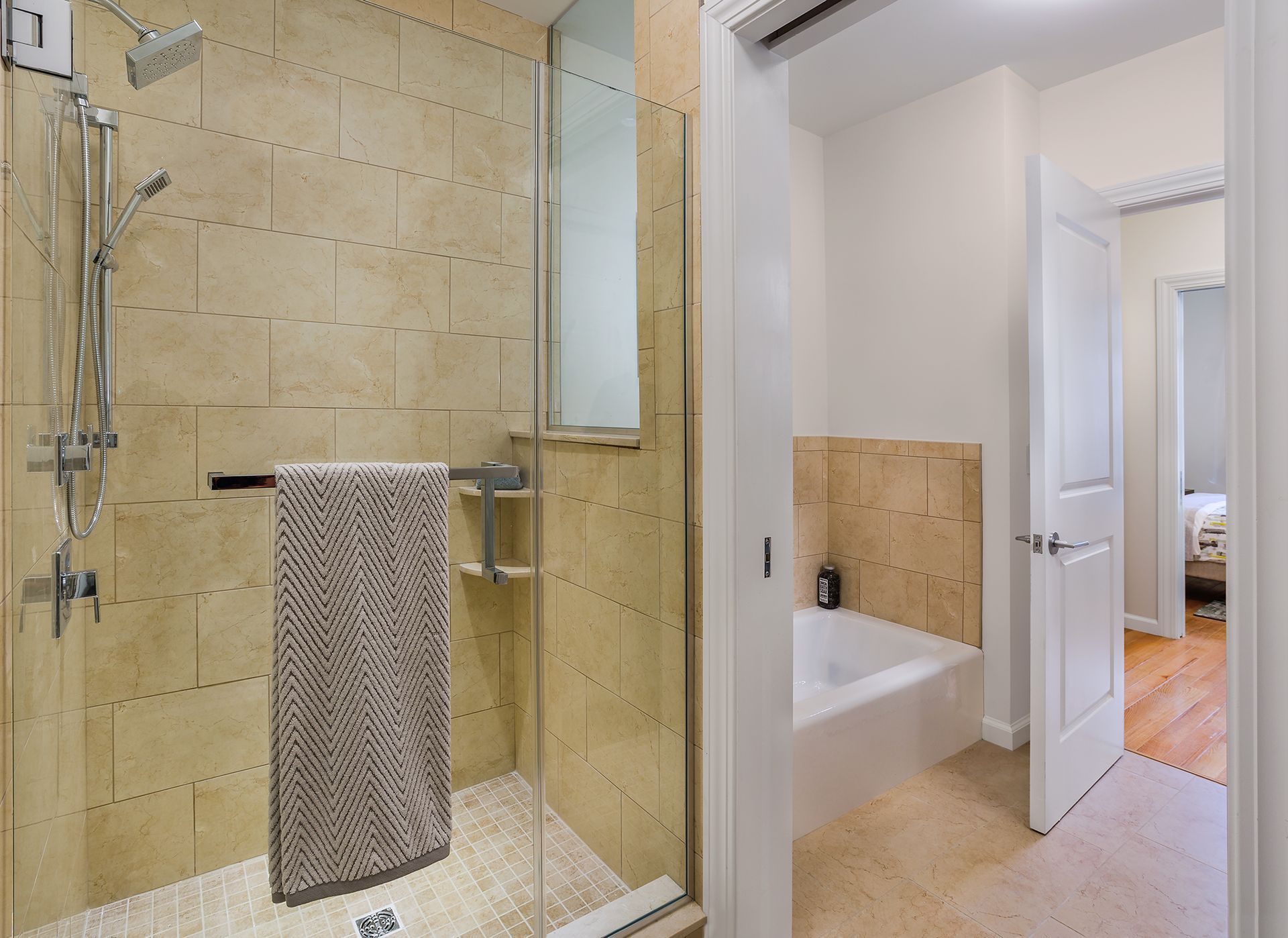 Bathroom with glass shower, towel, and tub. Beige tiled walls and flooring with white door.