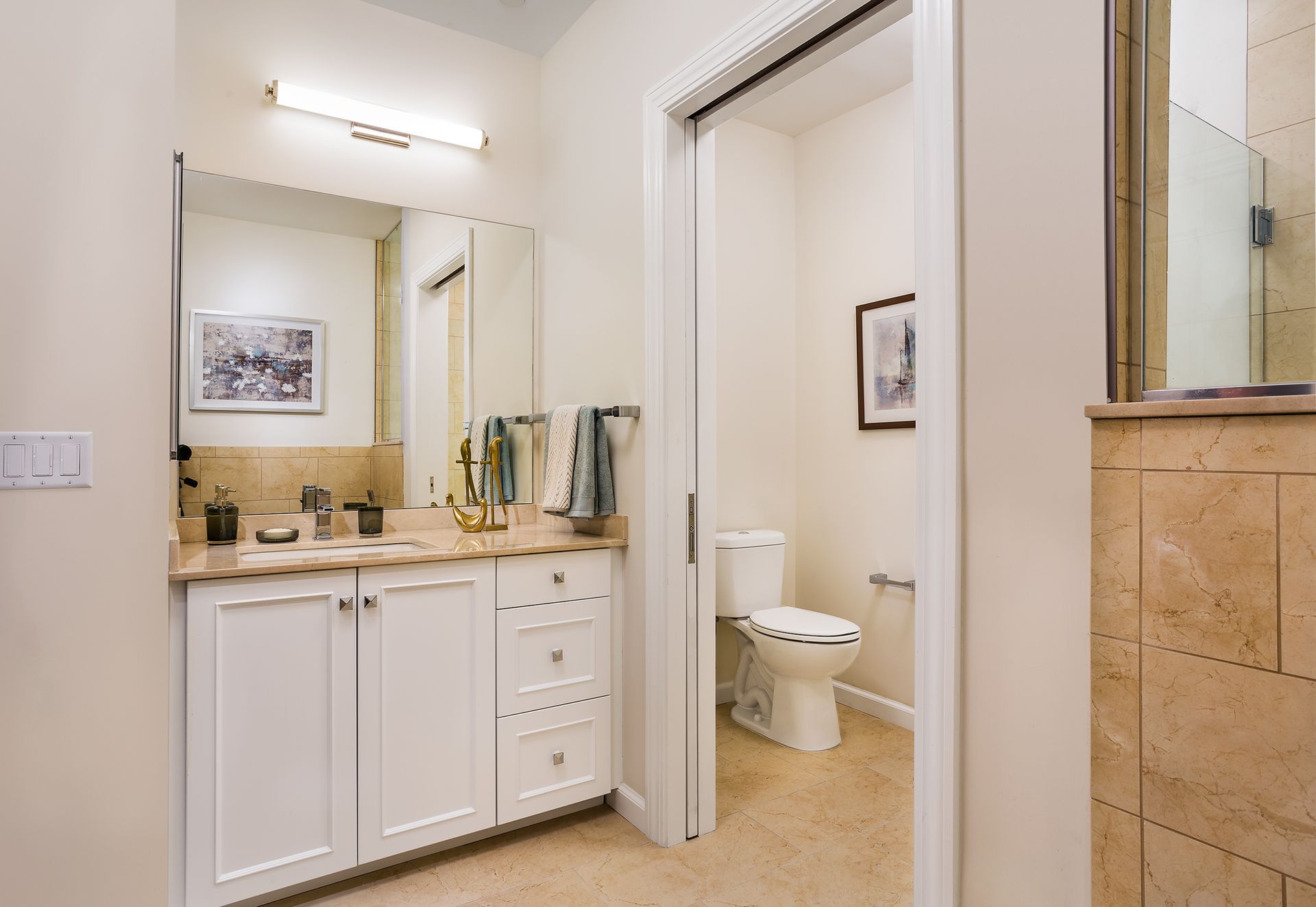Bathroom with white vanity, toilet in separate room, beige walls and tile, open door.
