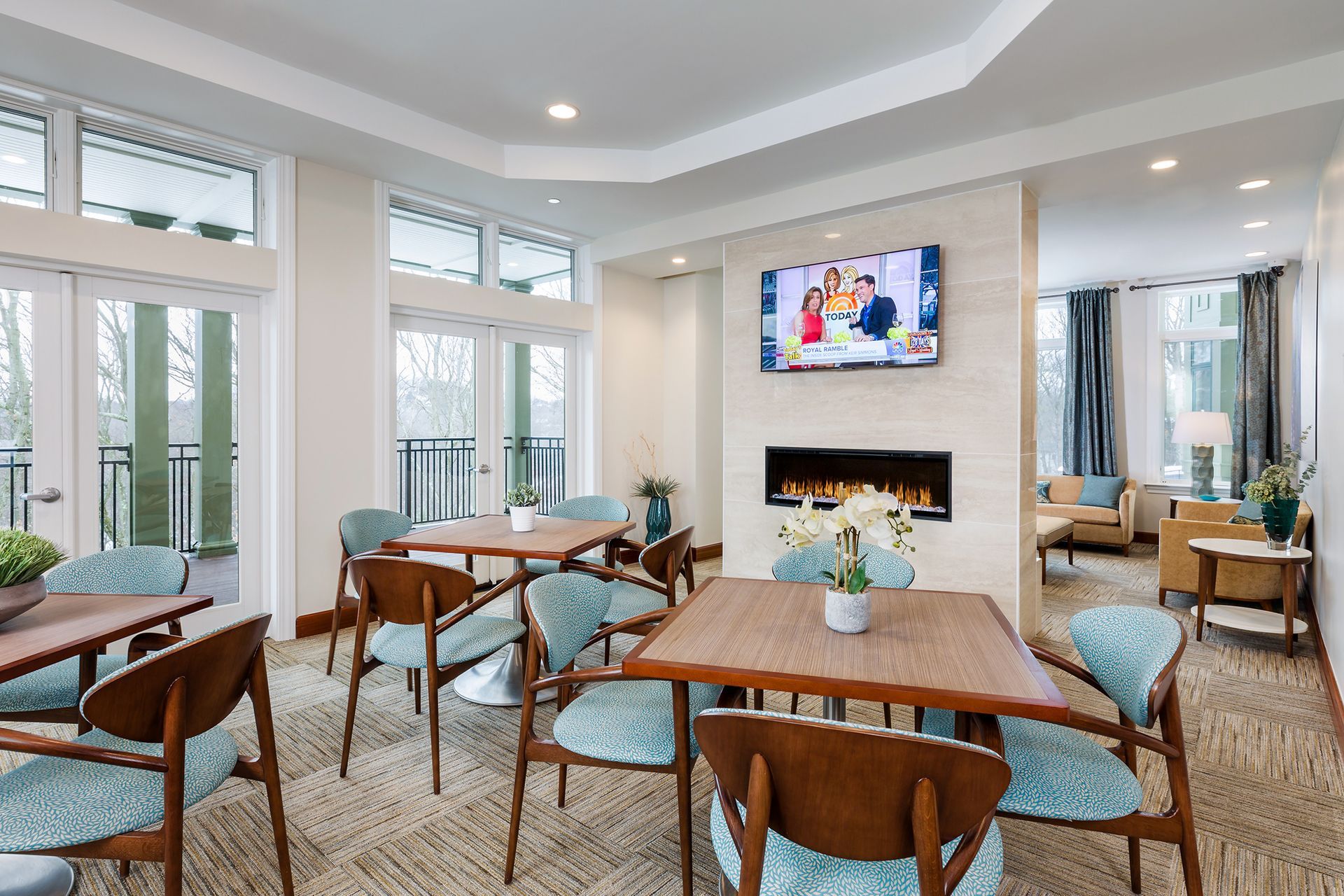 Cozy lounge with tables, chairs, fireplace, TV, and view of outdoor balcony.