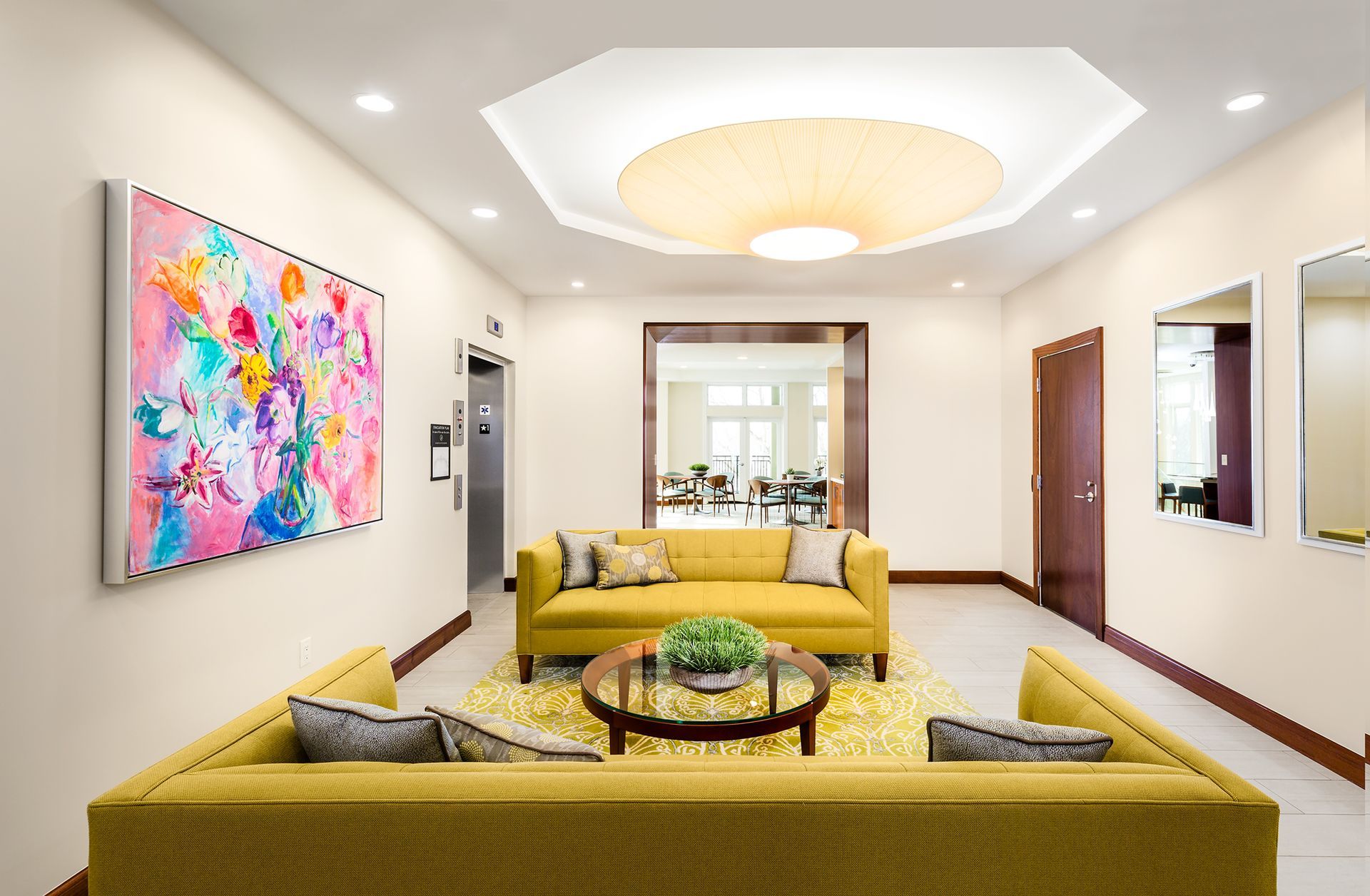 Lobby with yellow sofas, abstract art, round coffee table, a rug, elevator, mirrors, and doorway to a dining area.