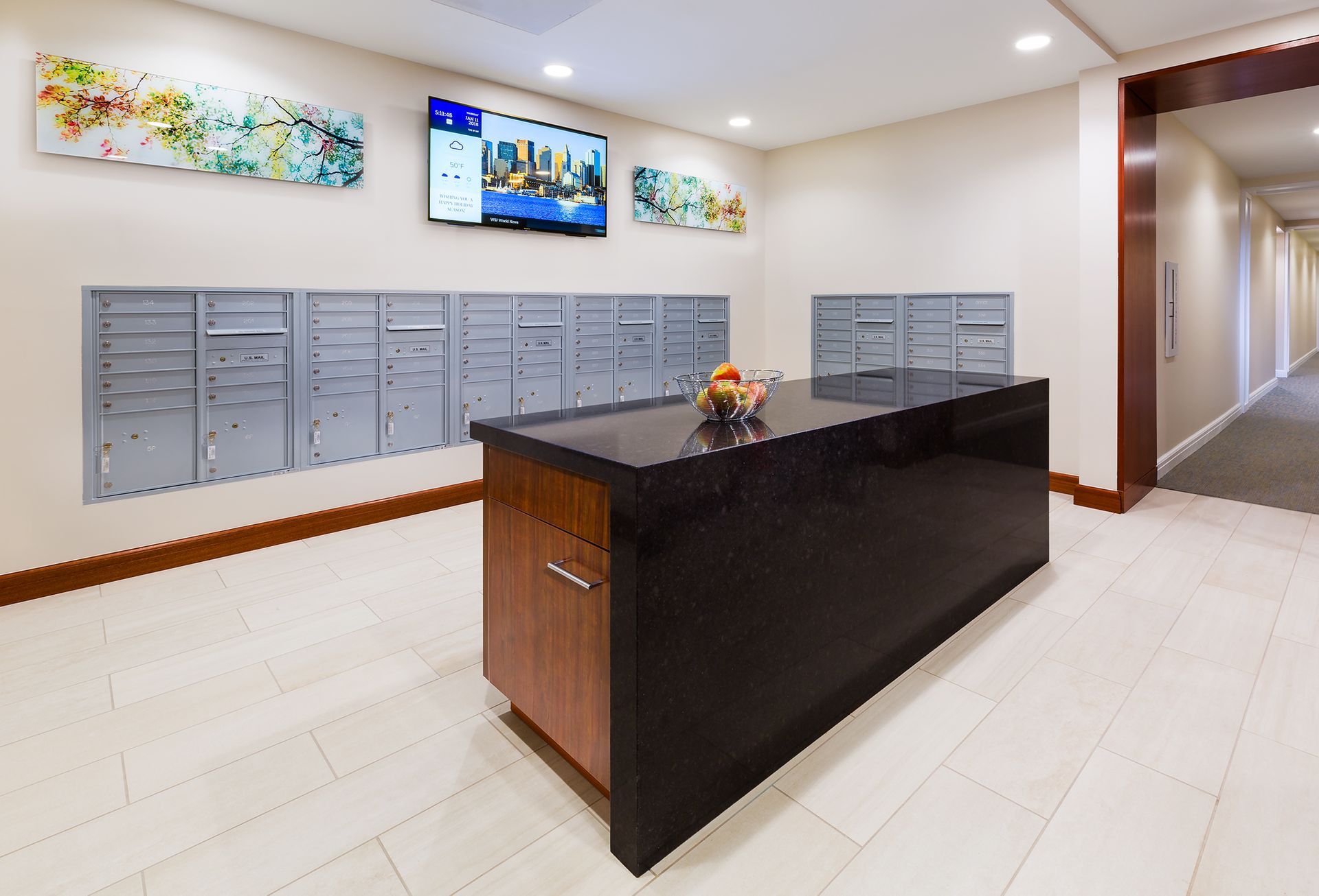 Lobby with mailboxes, countertop, TV, and artwork.  Beige walls, white floors, and a brown hallway are visible.