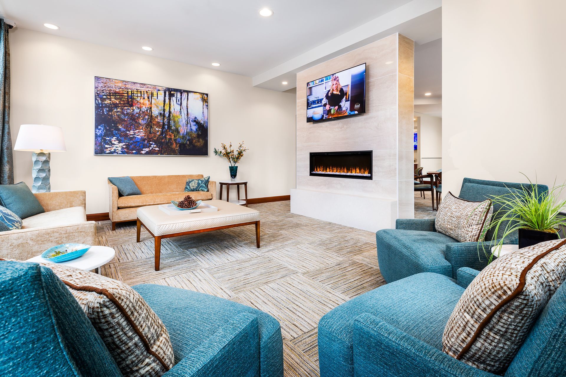 Cozy living room with teal armchairs, tan sofa, fireplace, and artwork. Neutral carpet and soft lighting.