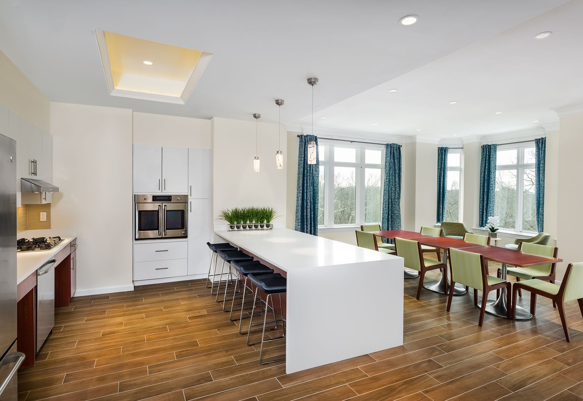 Modern kitchen with white island and cabinets, wooden floor, large windows with blue curtains, and long table with chairs.