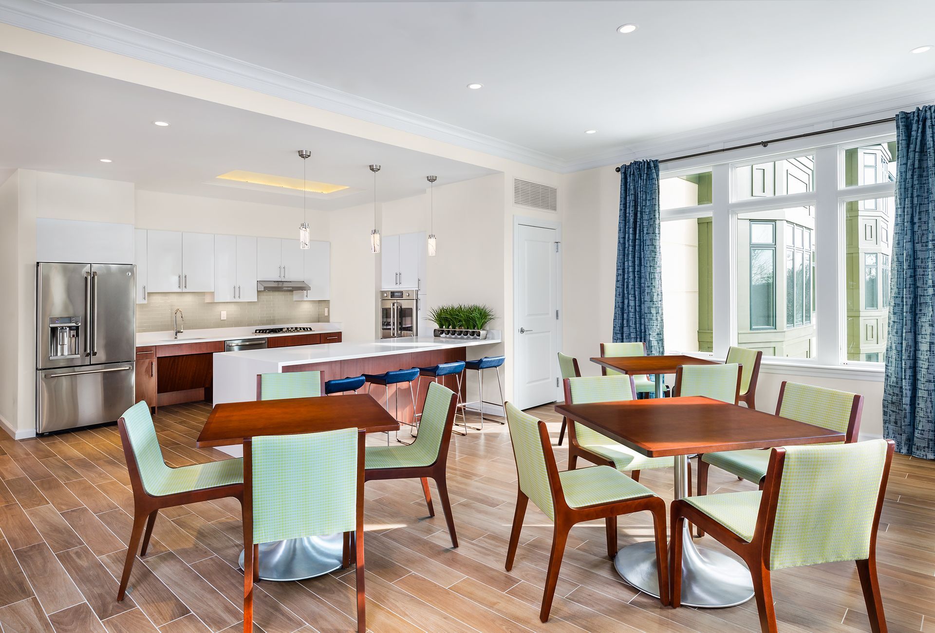Dining area with tables, chairs, and open kitchen; window with blue curtains, wood floor.