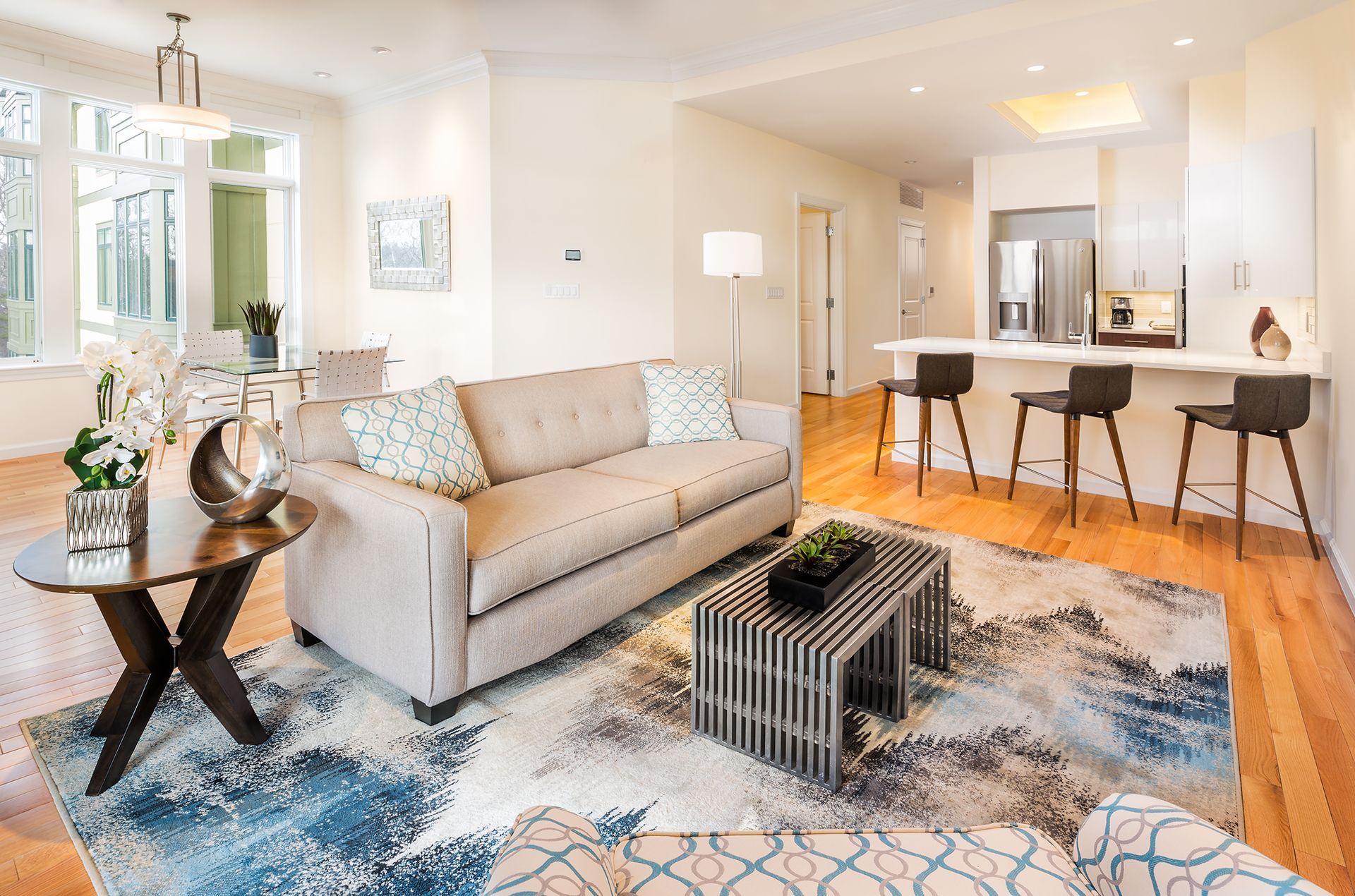 Living room with light-colored walls, sofa, patterned rug, kitchen with bar stools, and large windows.
