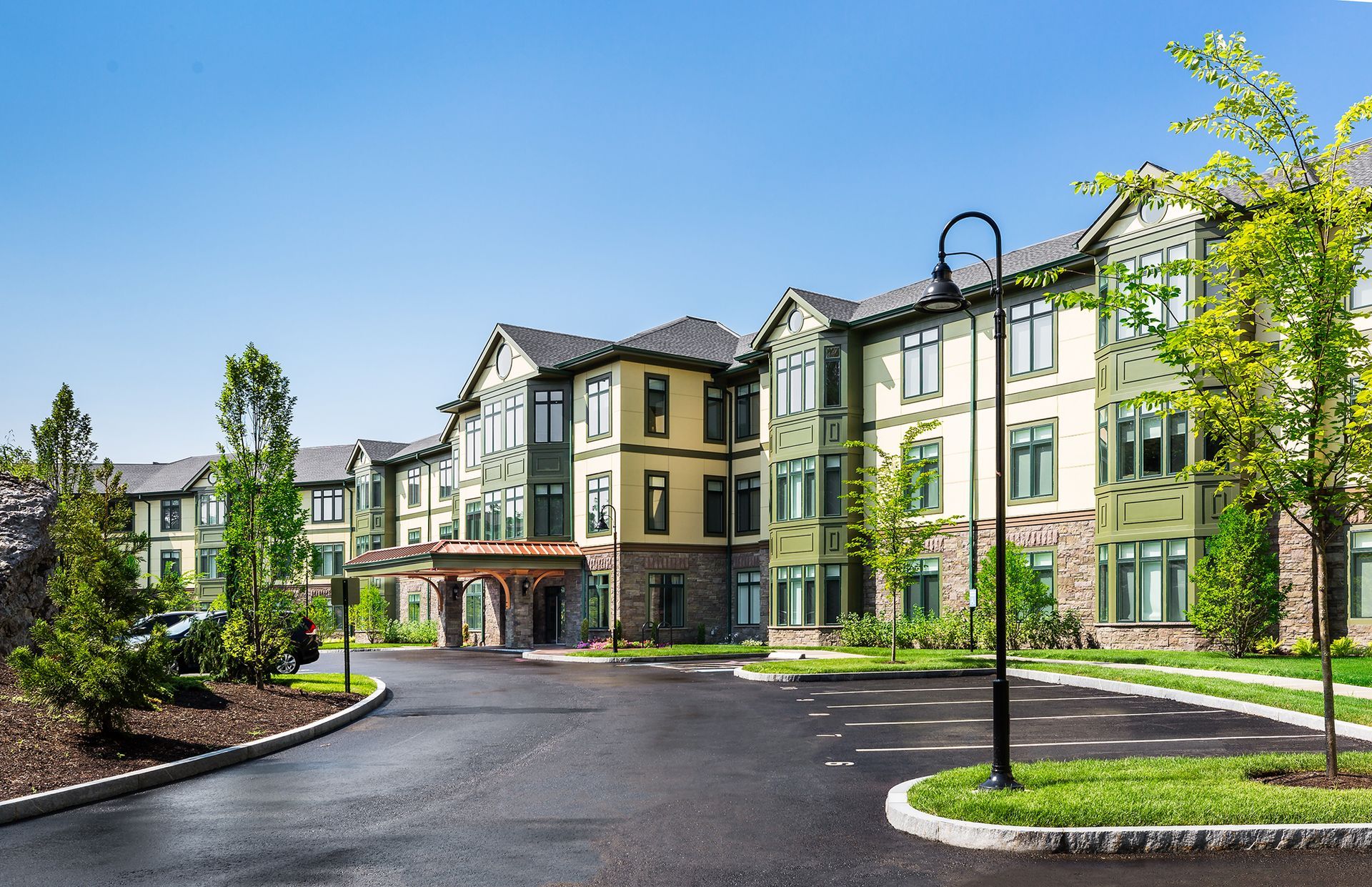 Exterior view of a three-story assisted living facility with asphalt drive. Green and tan building, blue sky.