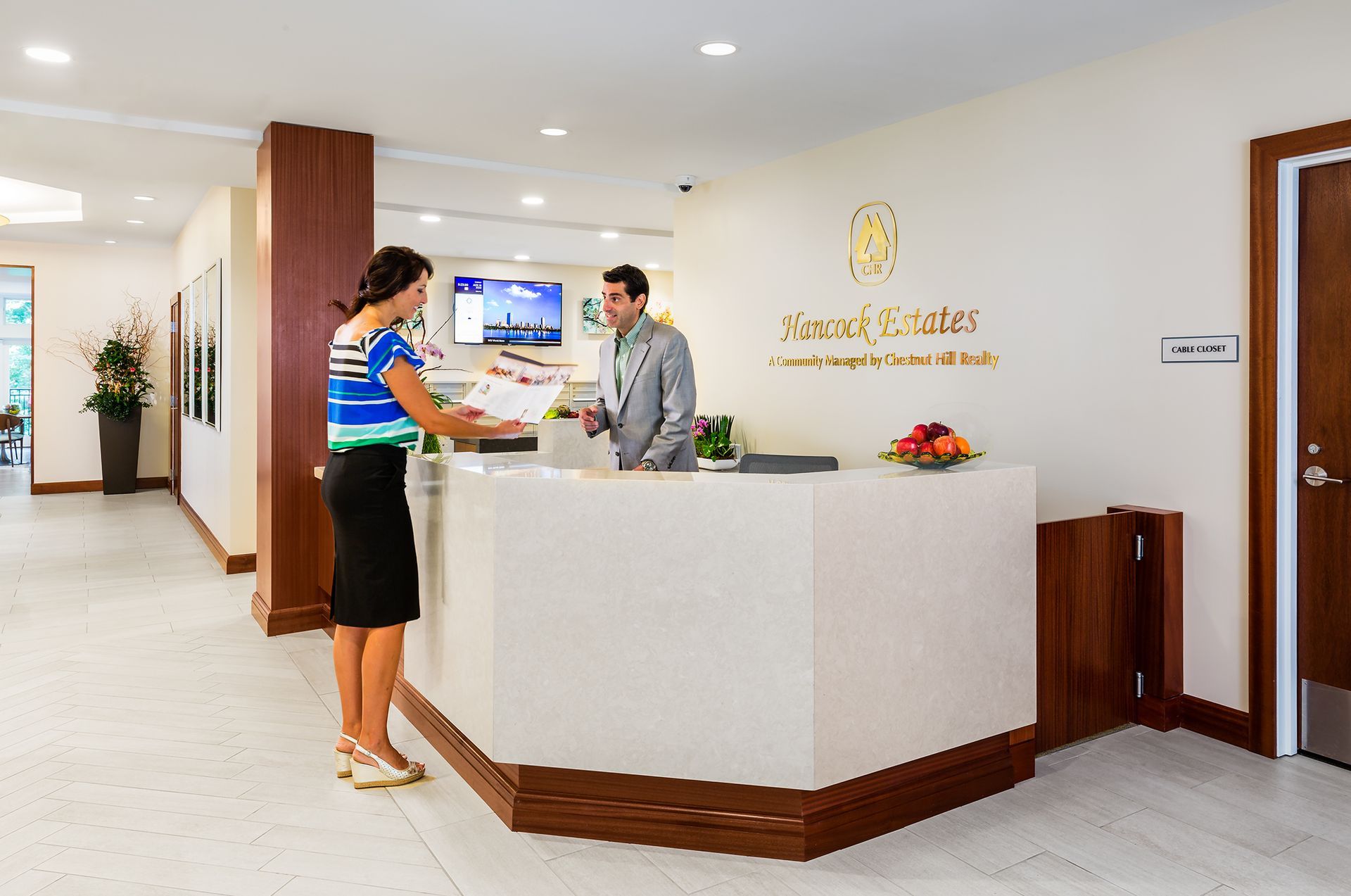Woman at reception desk with staff member; white counter, gold signage, bright interior.