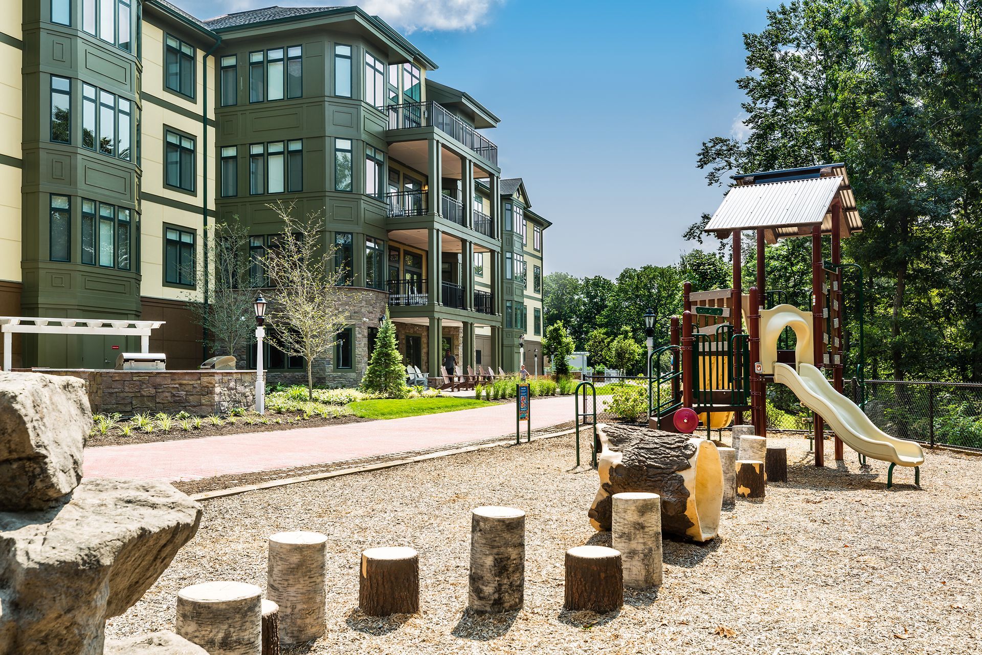 Apartment building with playground, path. Wooden posts, slide on pebble surface. Sunny day.