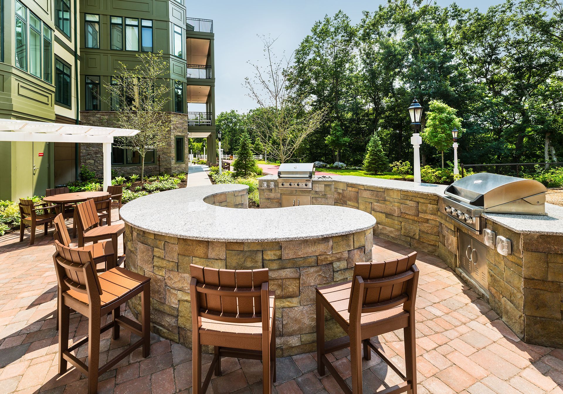 Outdoor grilling area with stone counters, built-in grills, and wooden bar chairs. Building in background.