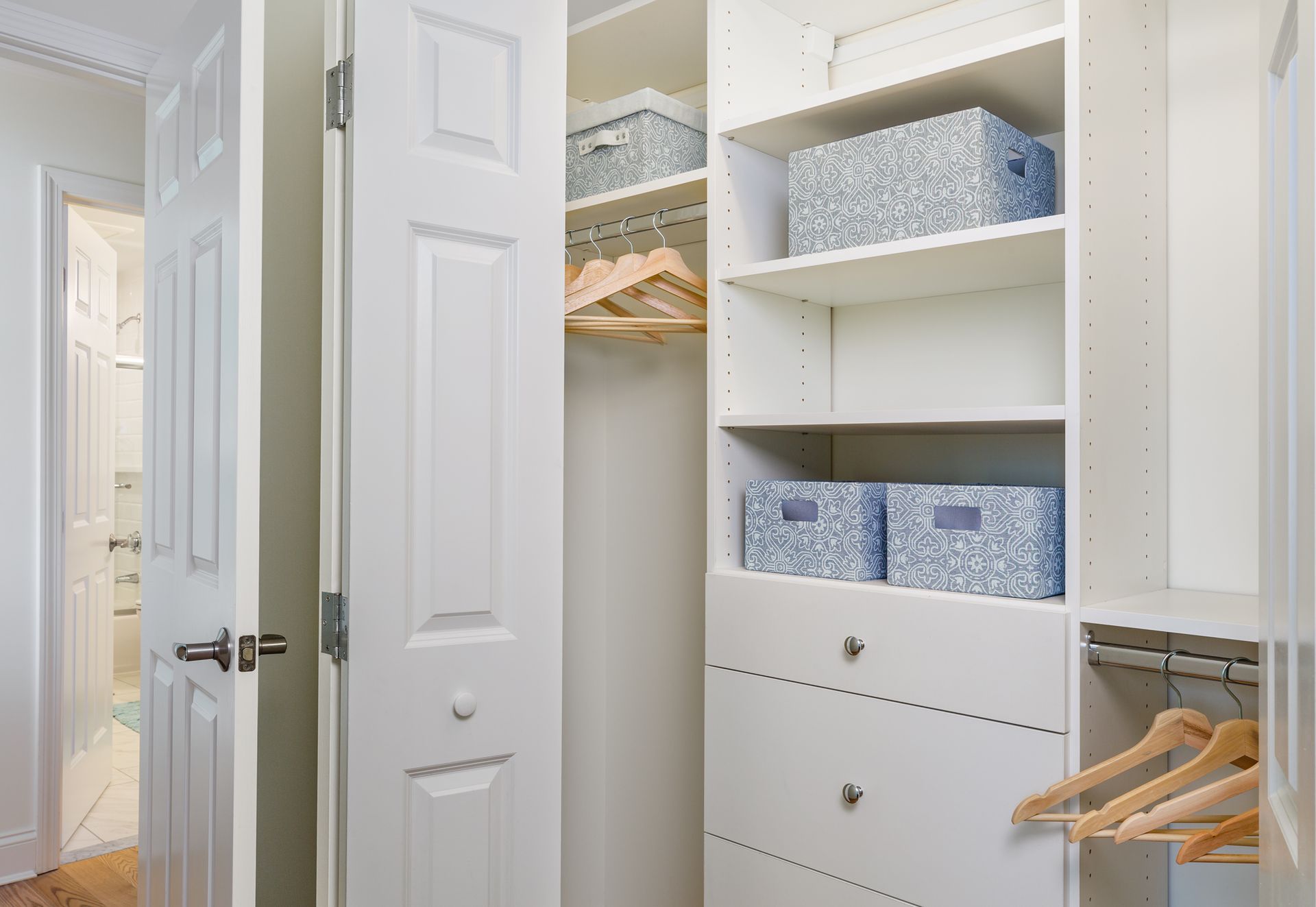 White organized closet with built-in shelving, drawers, and baskets. Open door reveals hallway and doorway.