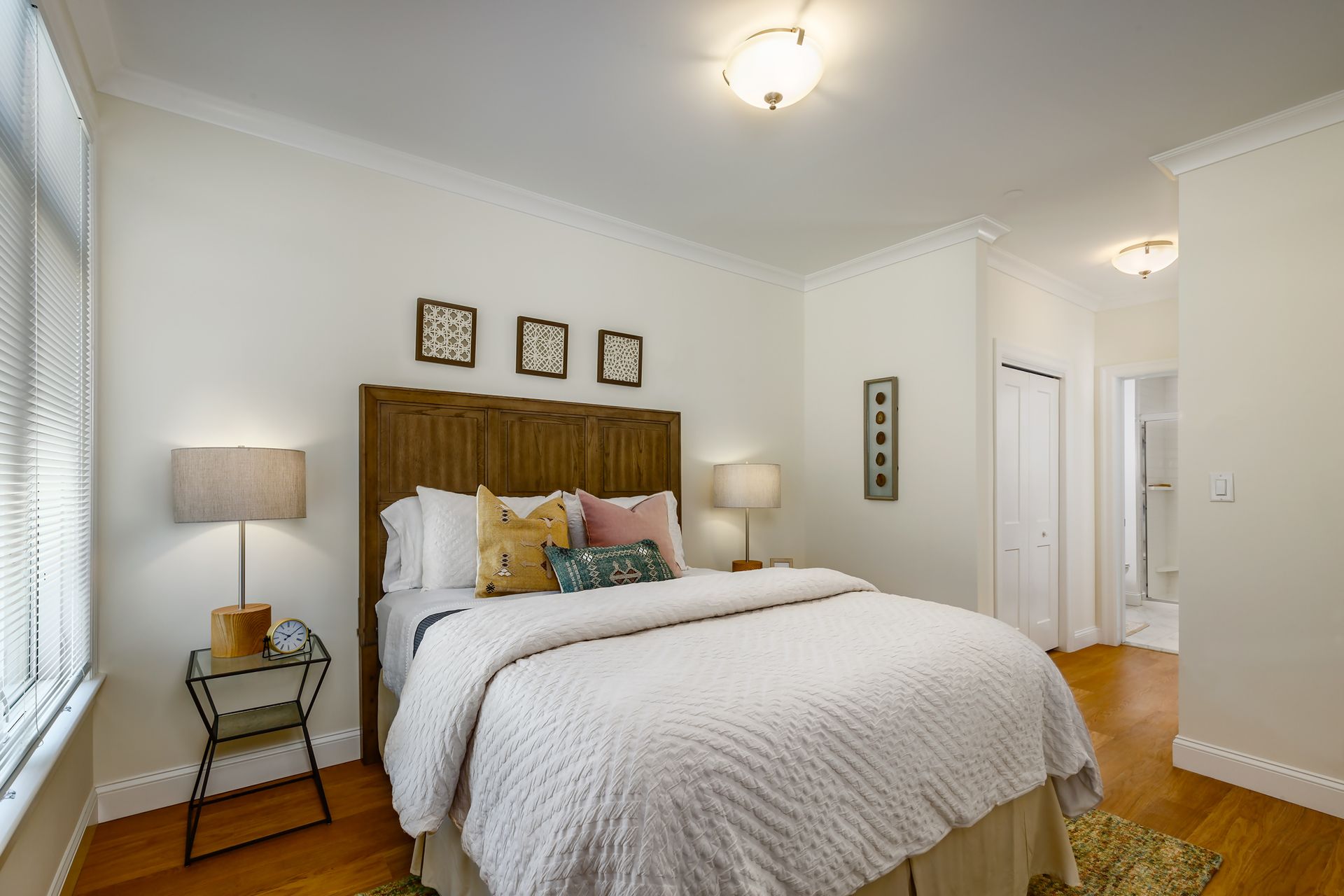 Bedroom with hardwood floors, white walls, bed with wooden headboard, side tables, and decorative pillows.