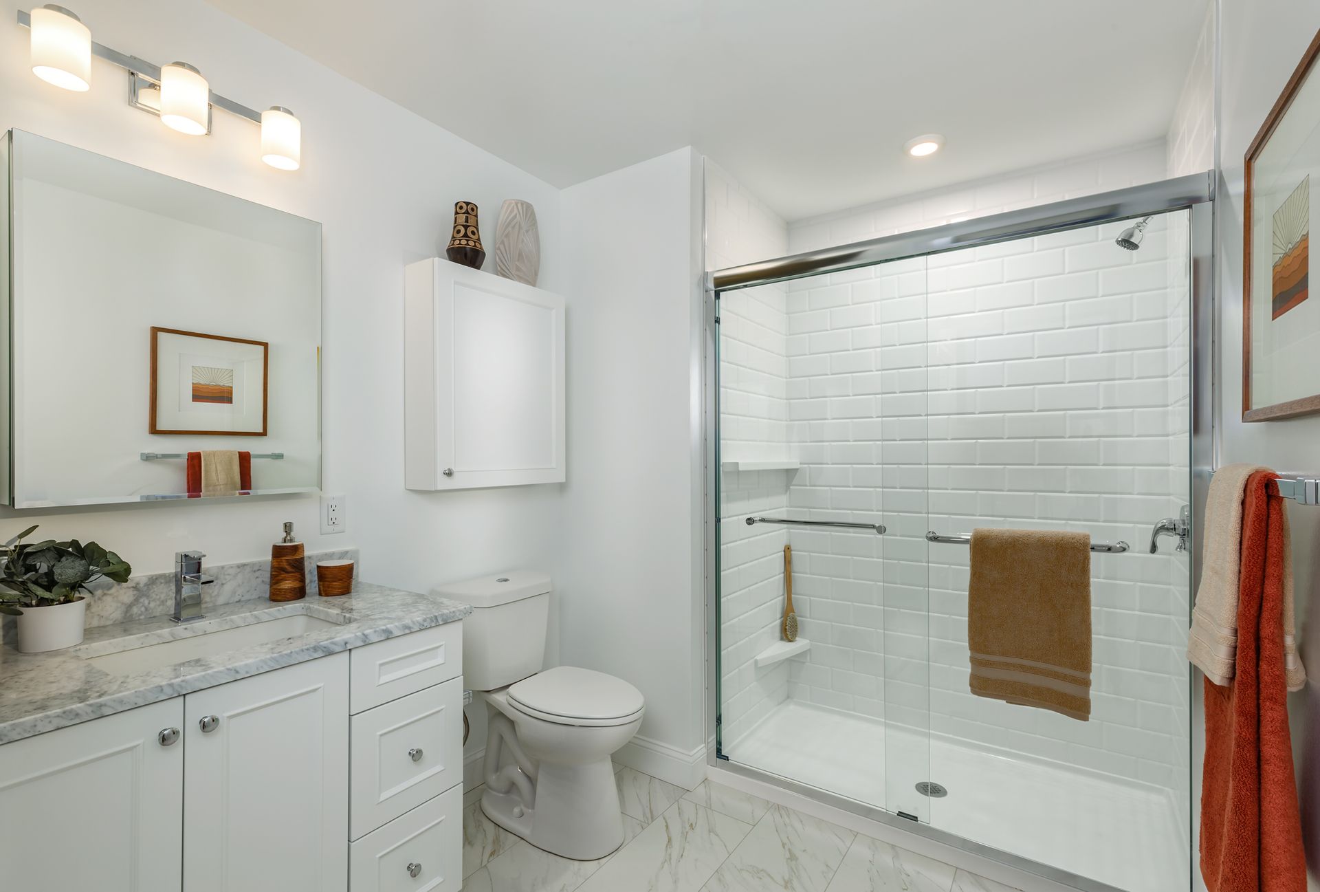 White bathroom with a shower, toilet, and vanity. Marble countertop, a mirror, and orange towels.