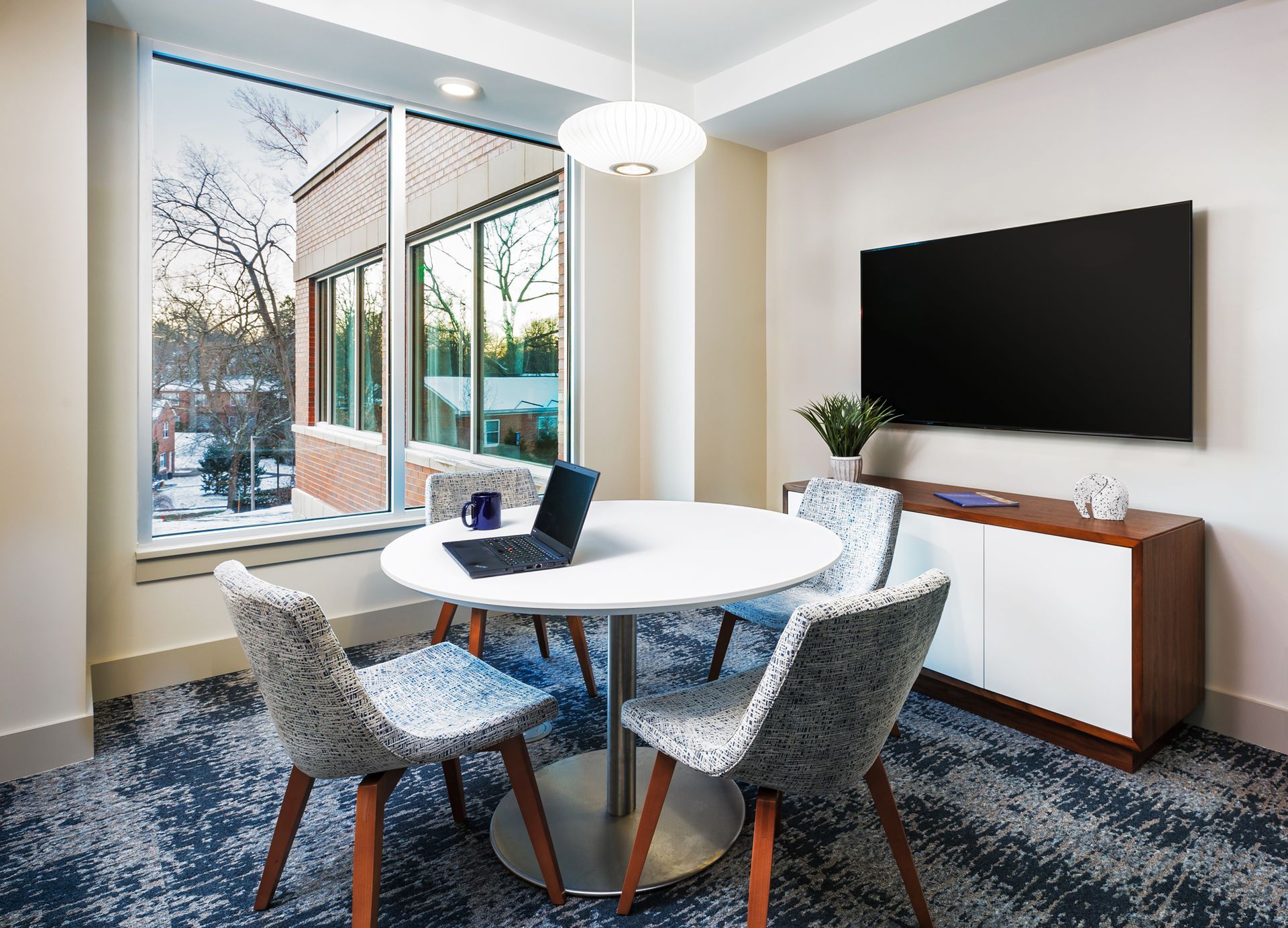 A modern office meeting room with a round table, chairs, and large TV. Large window overlooking trees.