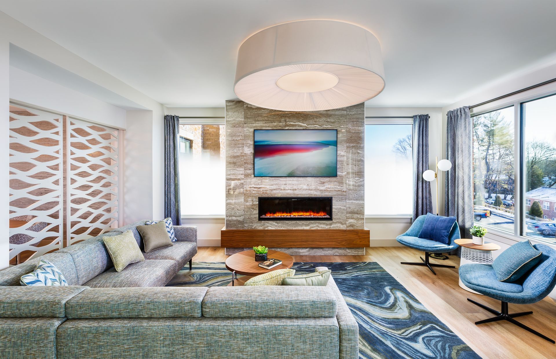 Modern living room with fireplace, gray sectional sofa, blue chairs, large windows, and a patterned rug.