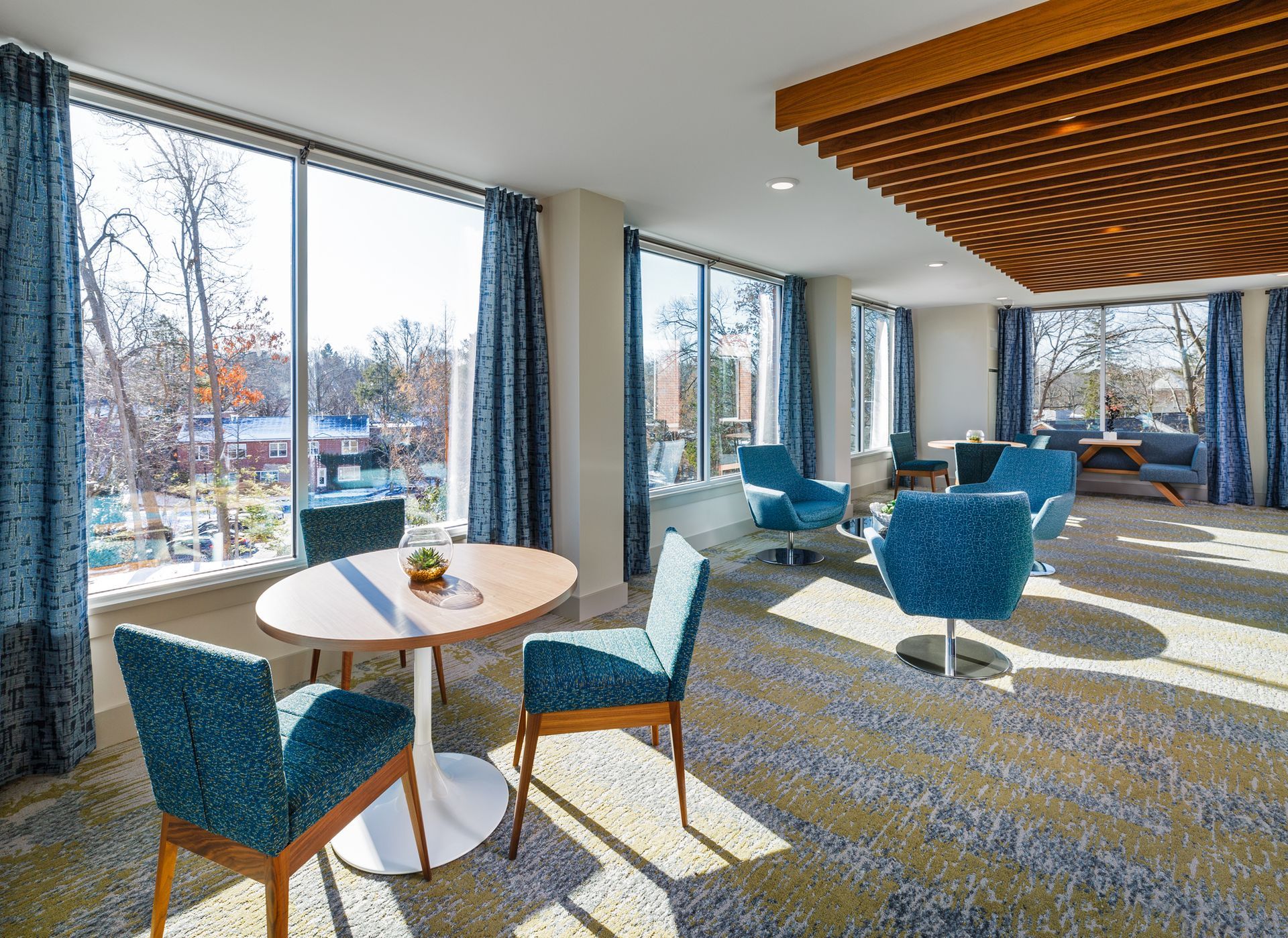 Bright room with large windows, round table, teal chairs. Open view, patterned carpet, wood ceiling.