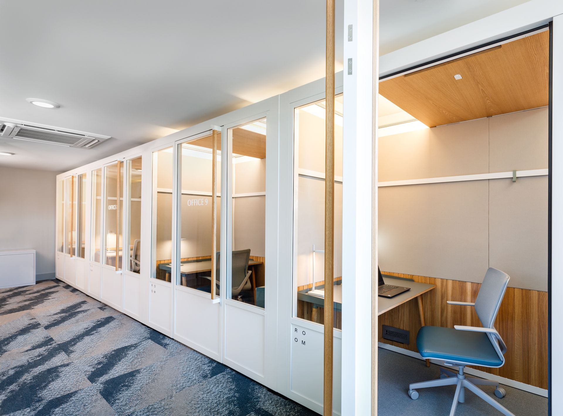 Row of white enclosed office booths with glass walls and work surfaces. Carpeted floor.