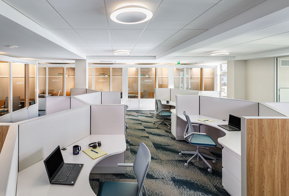Modern office cubicles with desks, chairs, and laptops. Overhead lighting, patterned carpet, and glass-walled rooms.