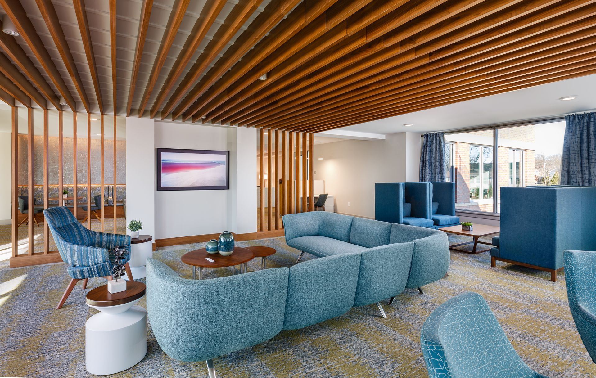 Lounge area with blue seating, wood accents, TV, and patterned carpet. Natural light and outdoor view.