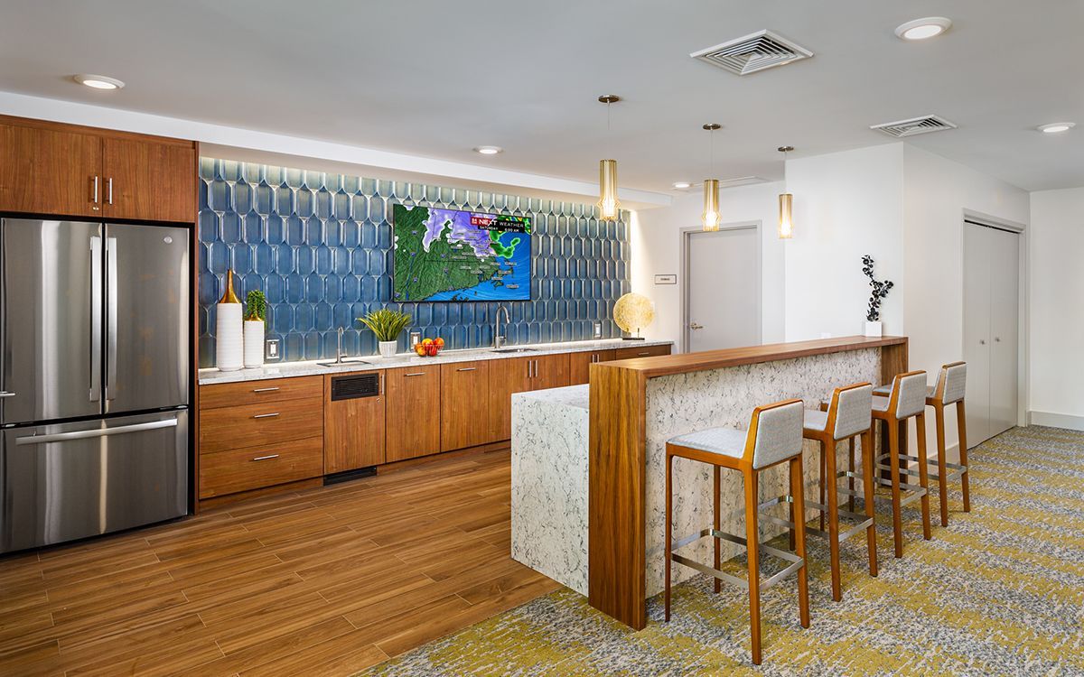 Modern kitchen area with wood cabinets, stainless steel refrigerator, blue tiled backsplash, and a bar with stools.