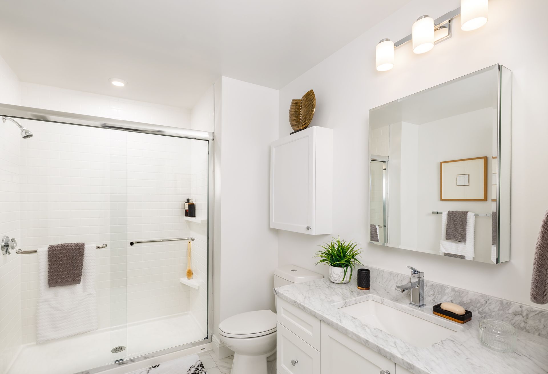 White bathroom with marble countertop, glass shower, and white cabinets.