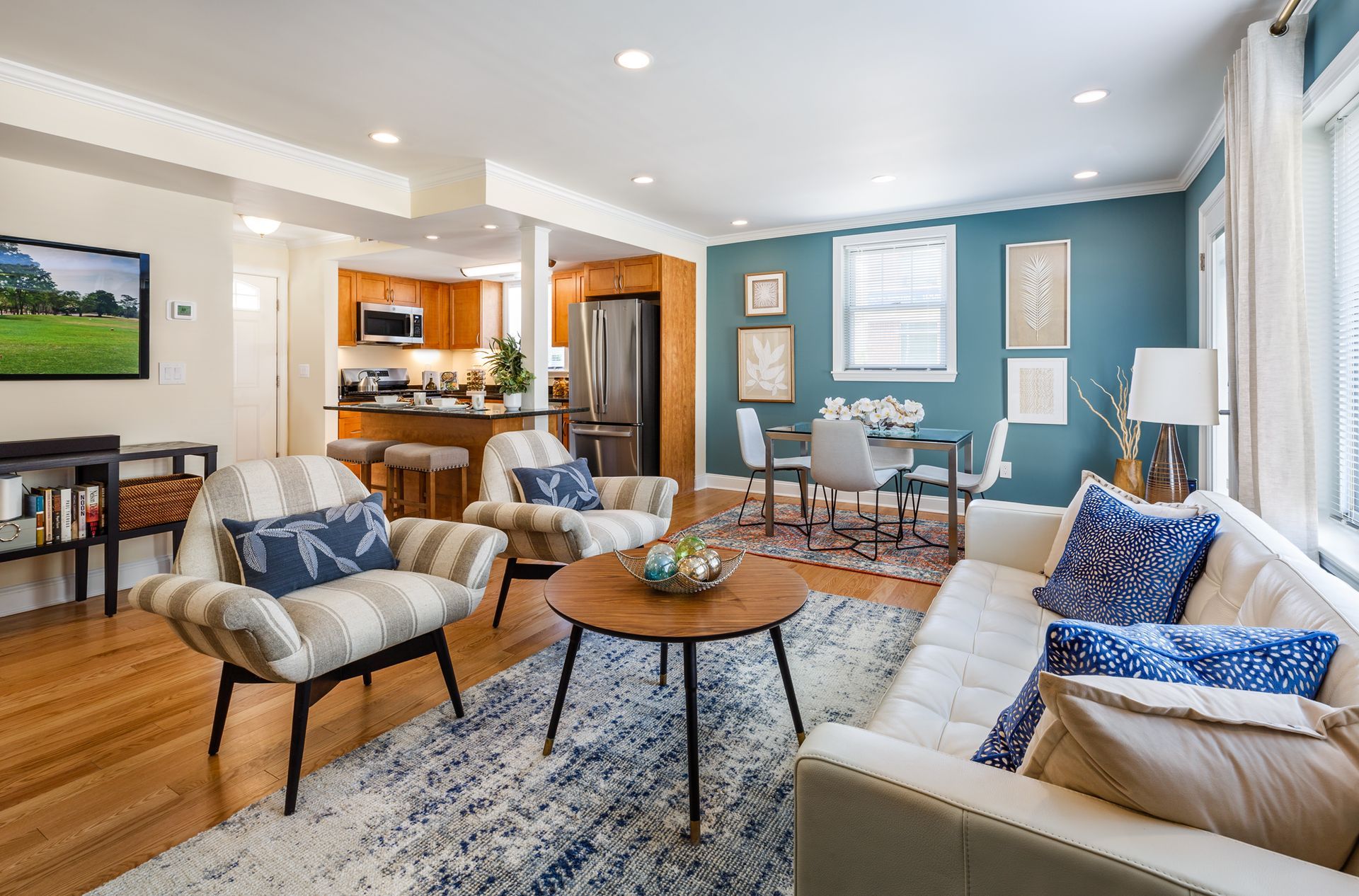 Bright living room with teal walls, beige sofa, patterned chairs, coffee table, and open kitchen view