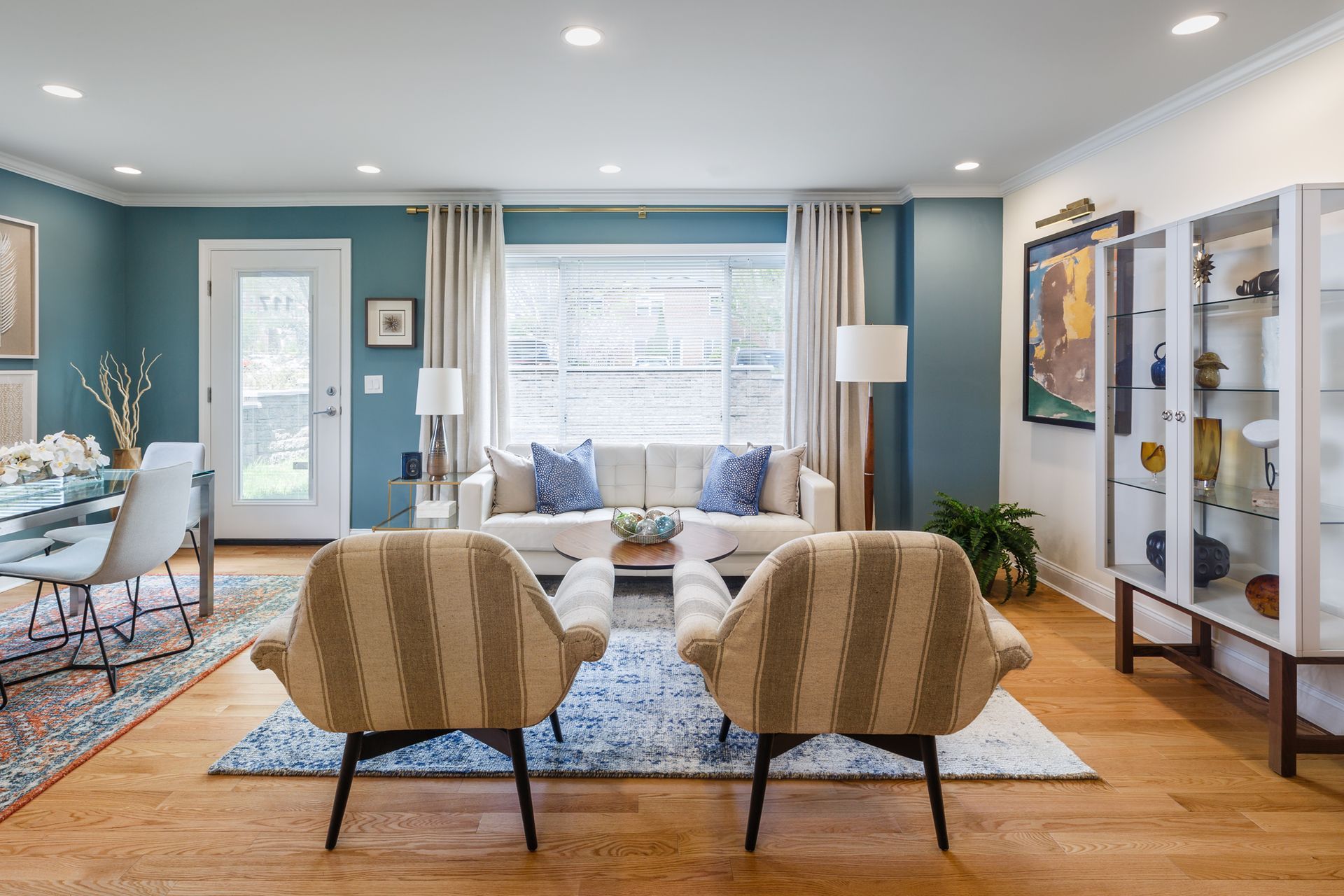 Bright living room with teal walls, tan chairs, sofa, patterned rug, and glass display cabinet.