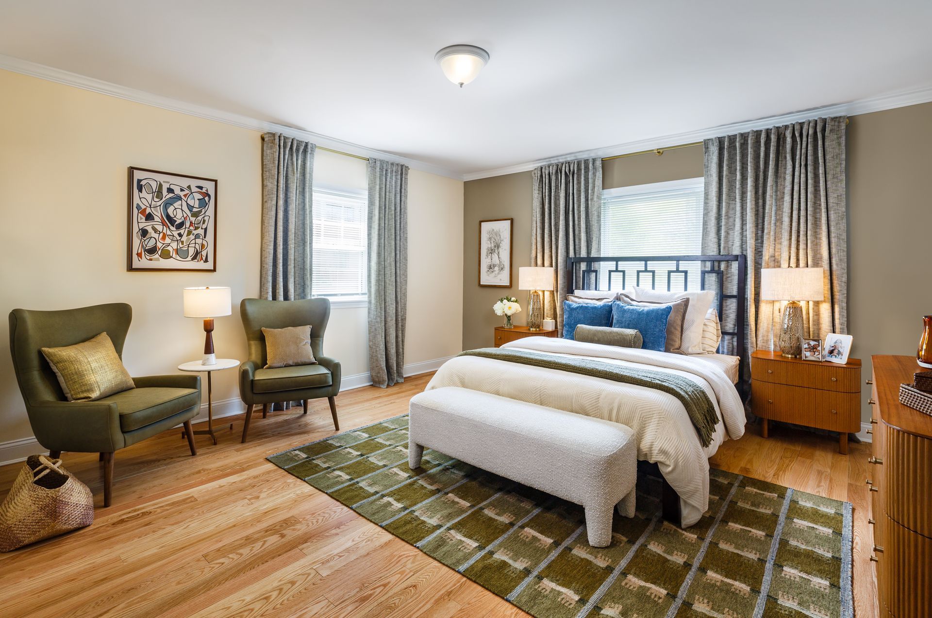 Cozy bedroom with a bed, bench, two chairs, nightstands, and patterned green rug beside large windows.