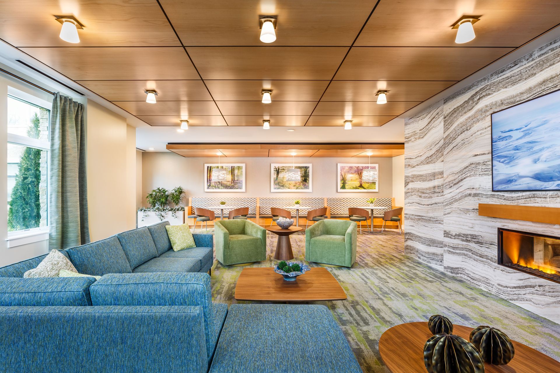 Cozy lounge with blue sectional, fireplace, and green armchairs. Neutral tones with wood and art.