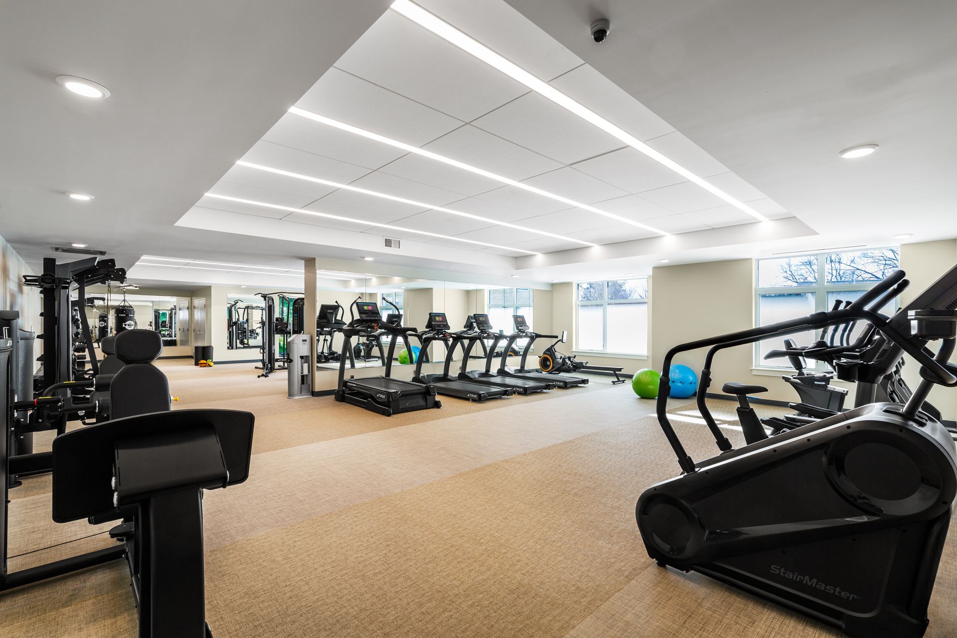 A modern fitness center featuring cardio machines, exercise balls, and neutral-toned carpeting under bright linear lights.