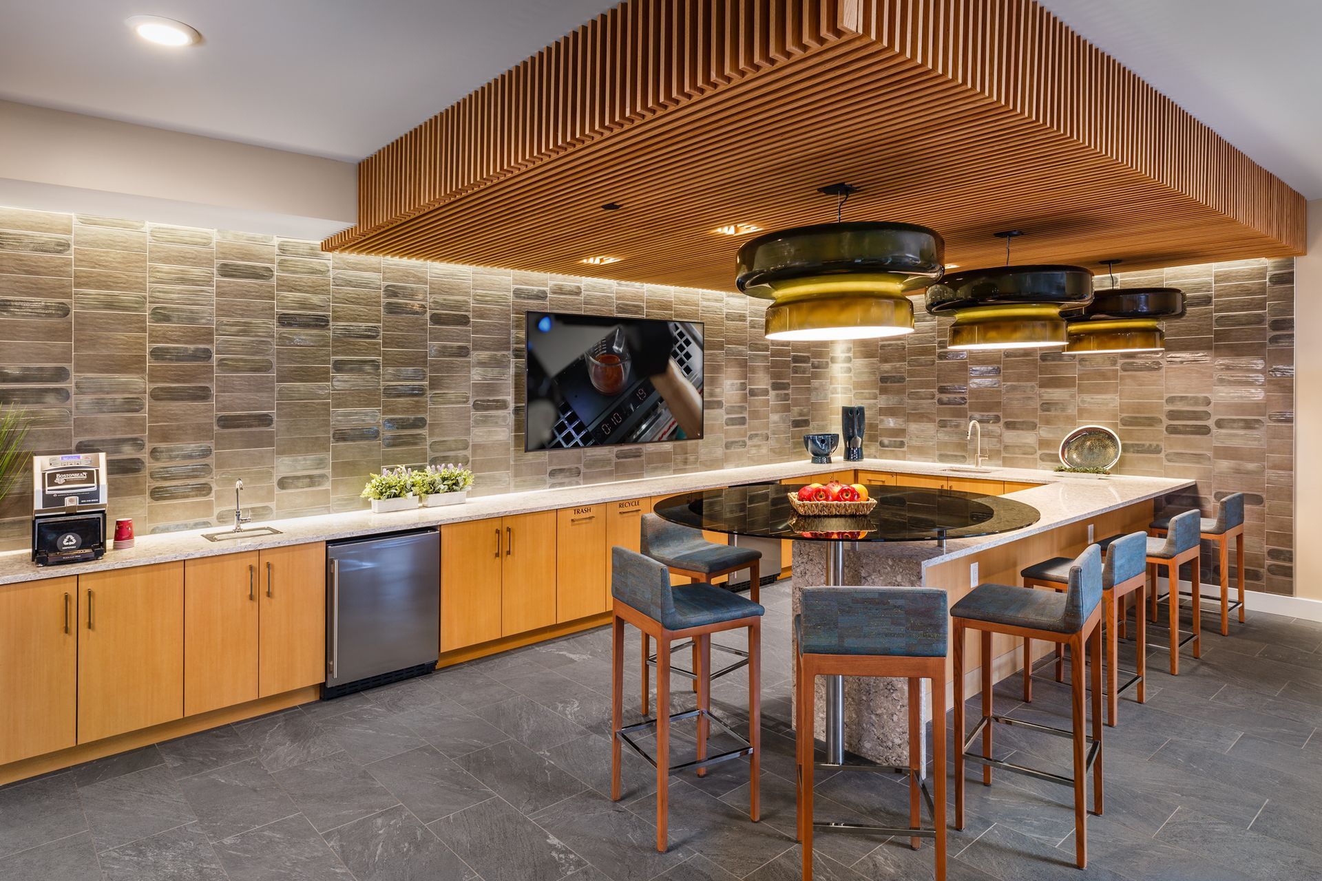 Modern kitchen area featuring light wood cabinets, stone wall, a large circular center island, and tiered wooden ceiling.