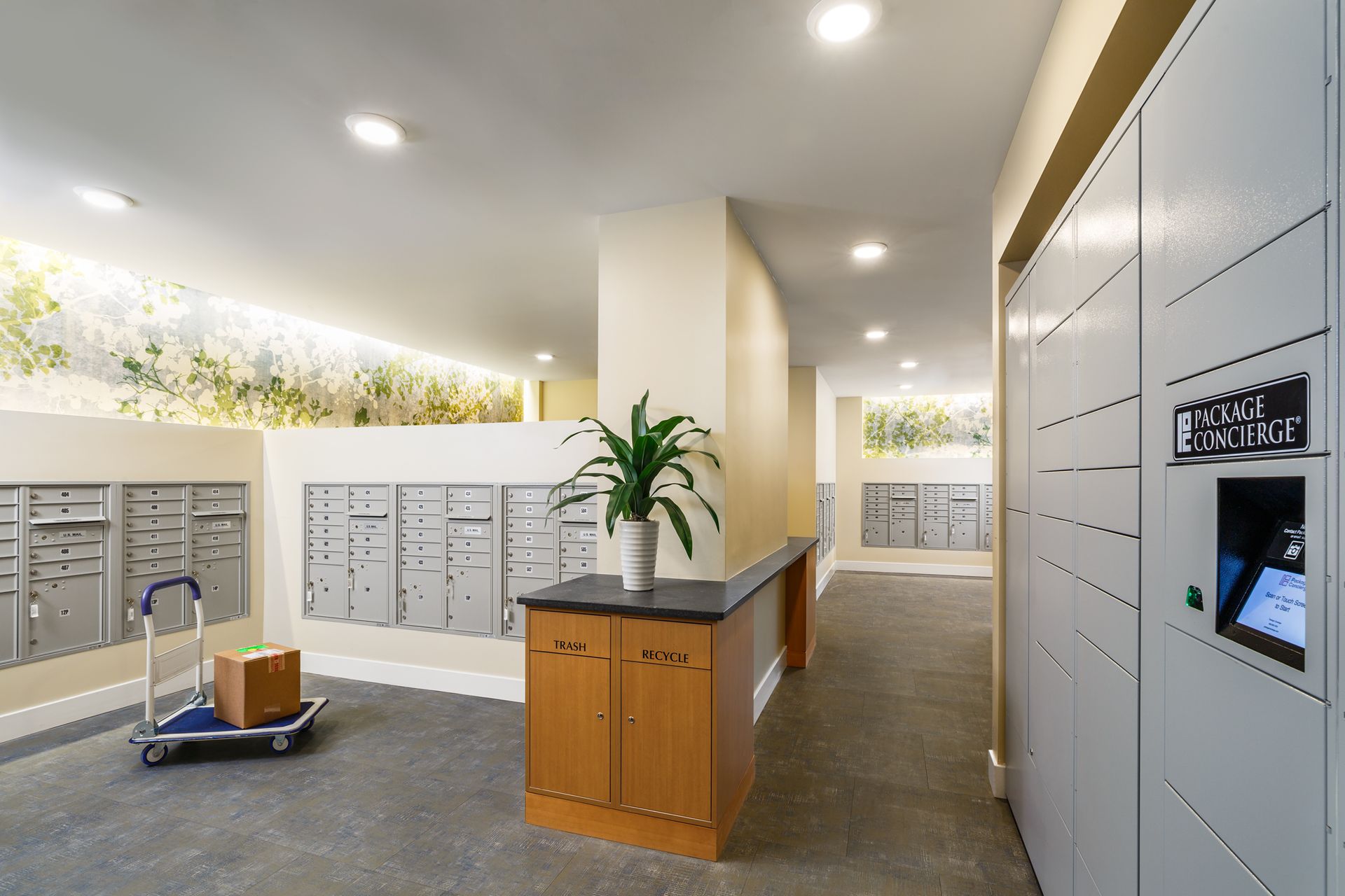 Lobby with mailboxes, package lockers, and a cart carrying a box. Neutral colors and overhead lighting.