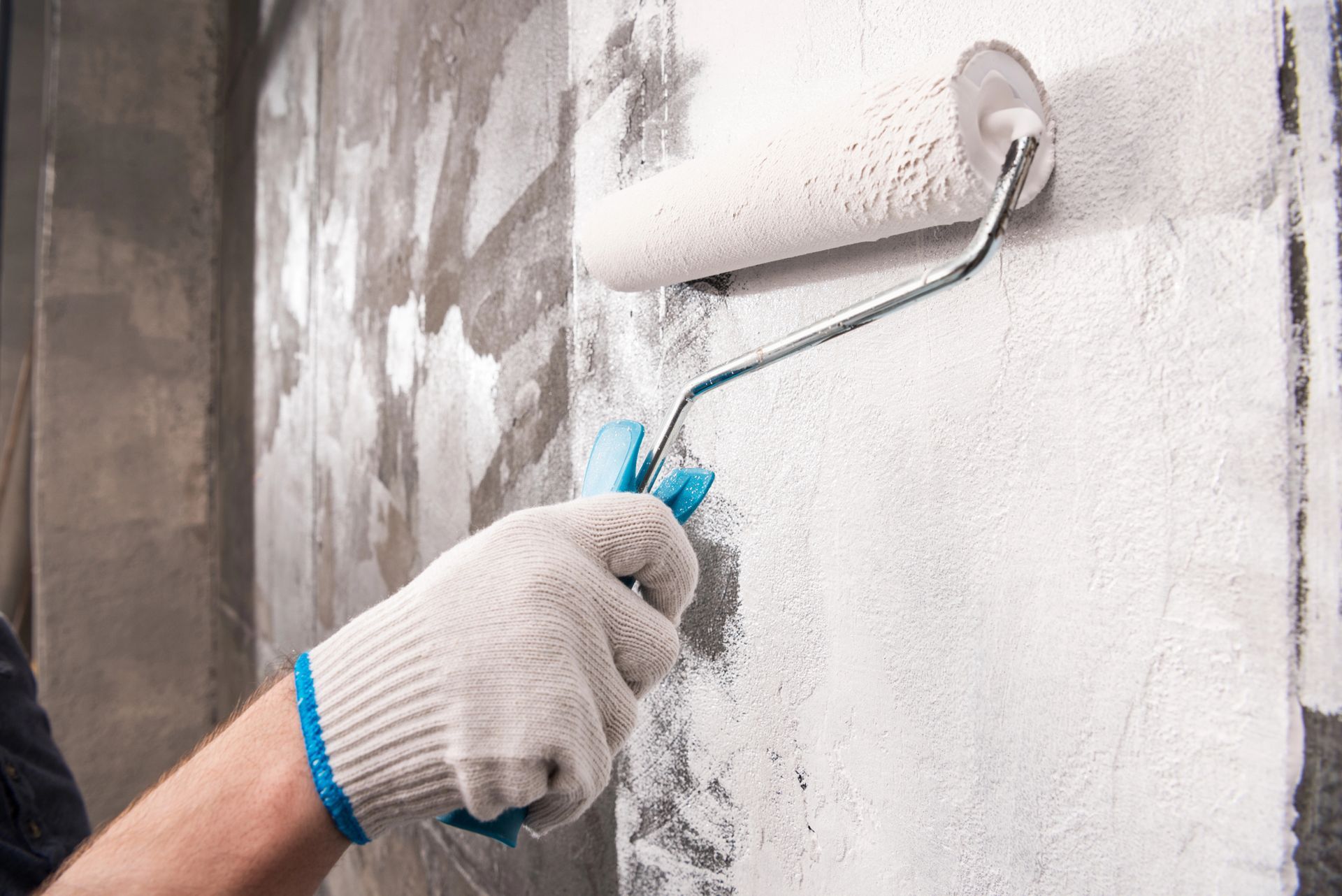Persona che indossa un guanto da lavoro e usa un rullo per dipingere su un muro di cemento, applicando vernice bianca.