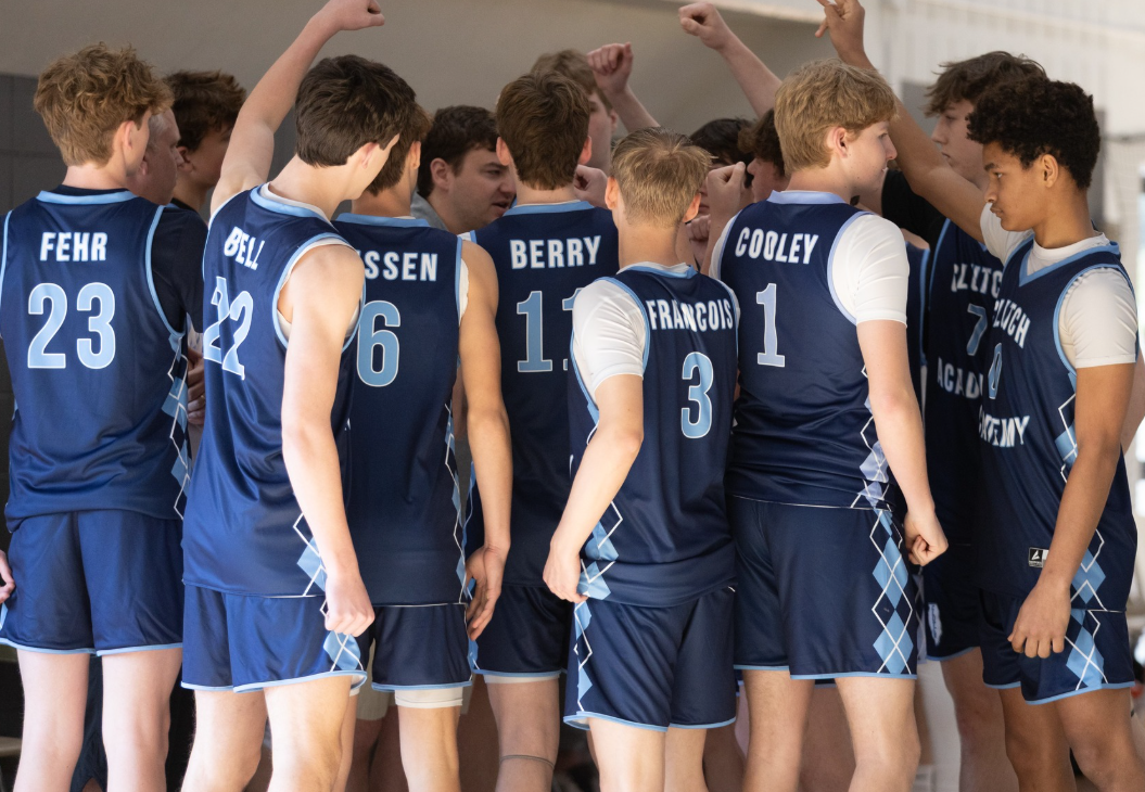 Volleyball team huddling indoors, wearing navy jerseys with numbers on their backs.