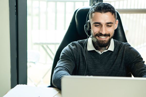 Man with headset smiles while working on a laptop in front of a window.