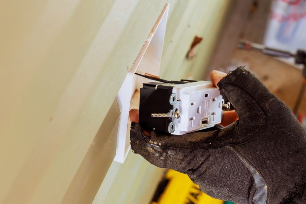 A gloved hand holds a new electrical outlet near a wall during installation.