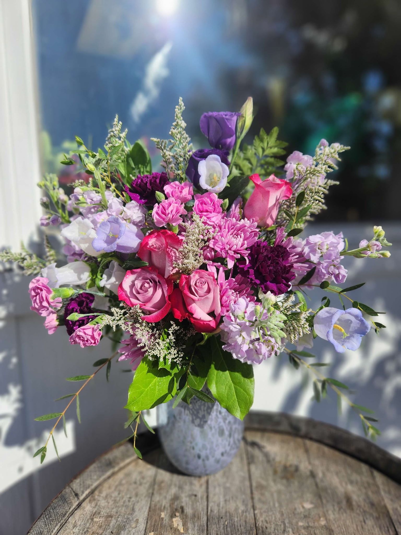 A vibrant bouquet of pink, purple, and white flowers in a speckled grey vase sitting on a wooden barrel.