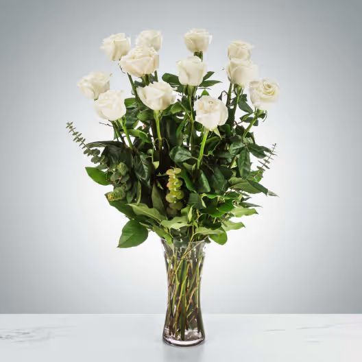 A bouquet of white roses with green foliage in a clear glass vase against a plain grey background.