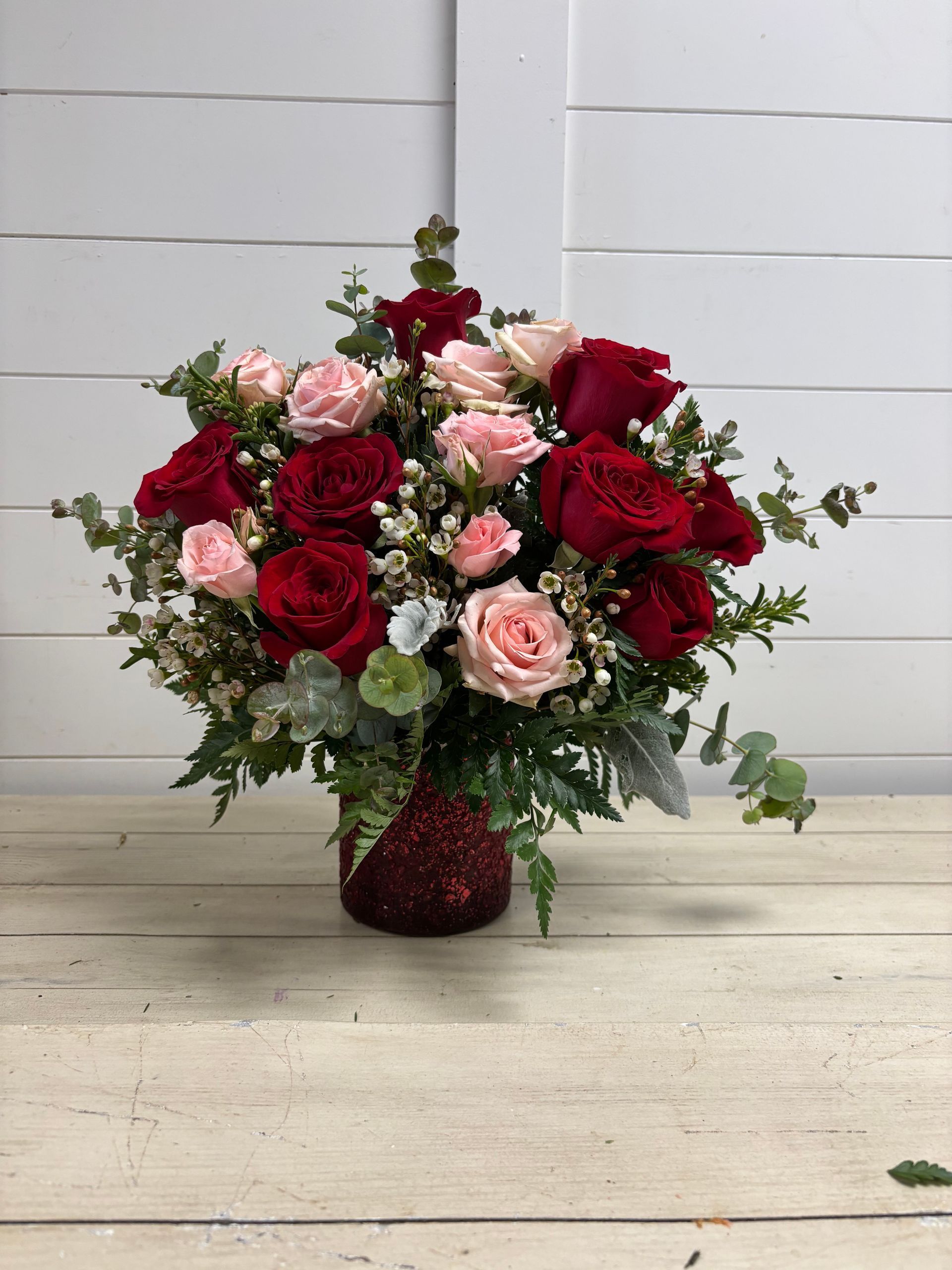 A bouquet of red and light pink roses with greenery in a textured red vase, set against a white horizontal slat wall.