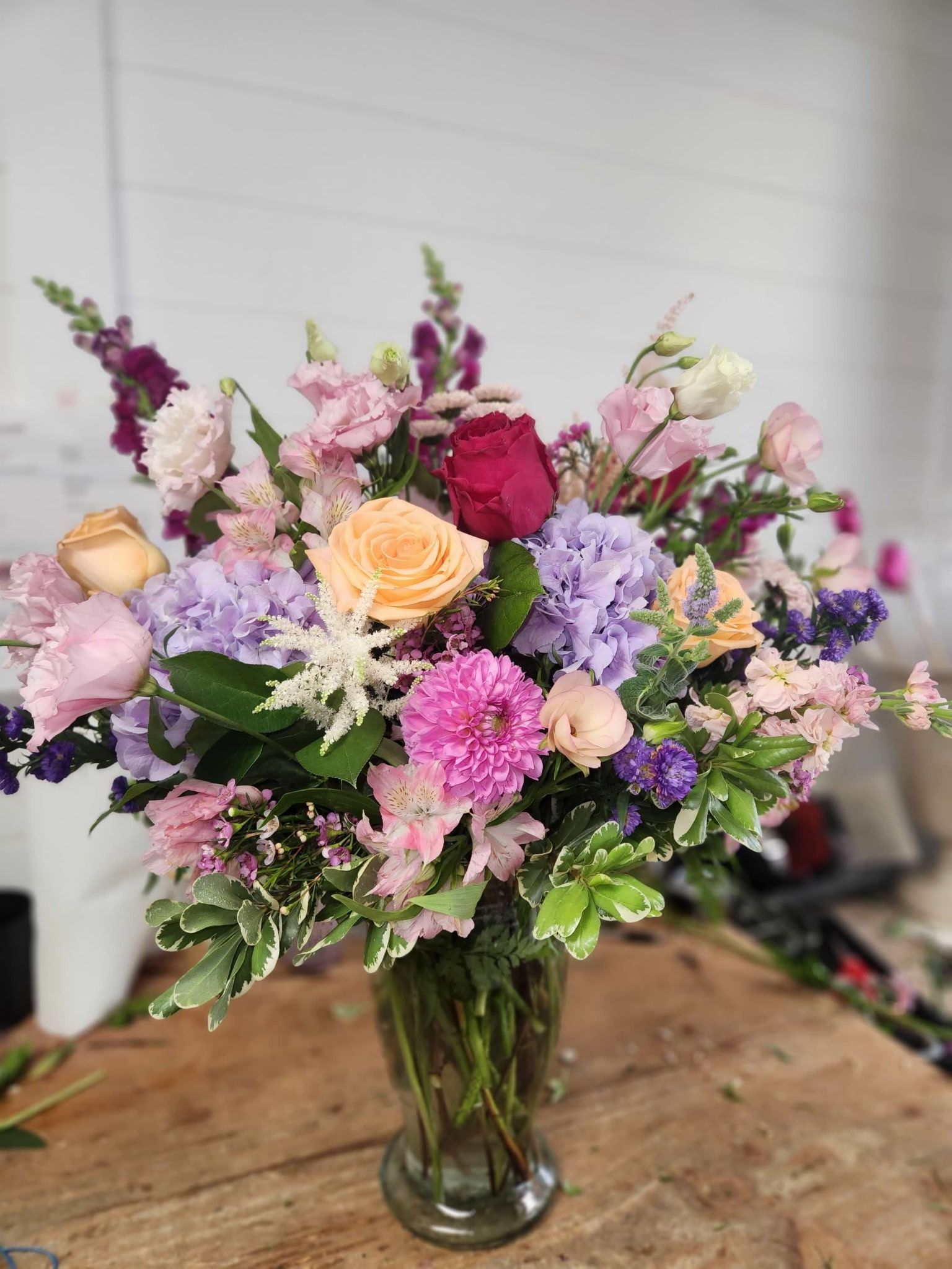 A glass vase filled with a vibrant, assorted floral arrangement featuring pink, purple, and peach flowers on a wood table.