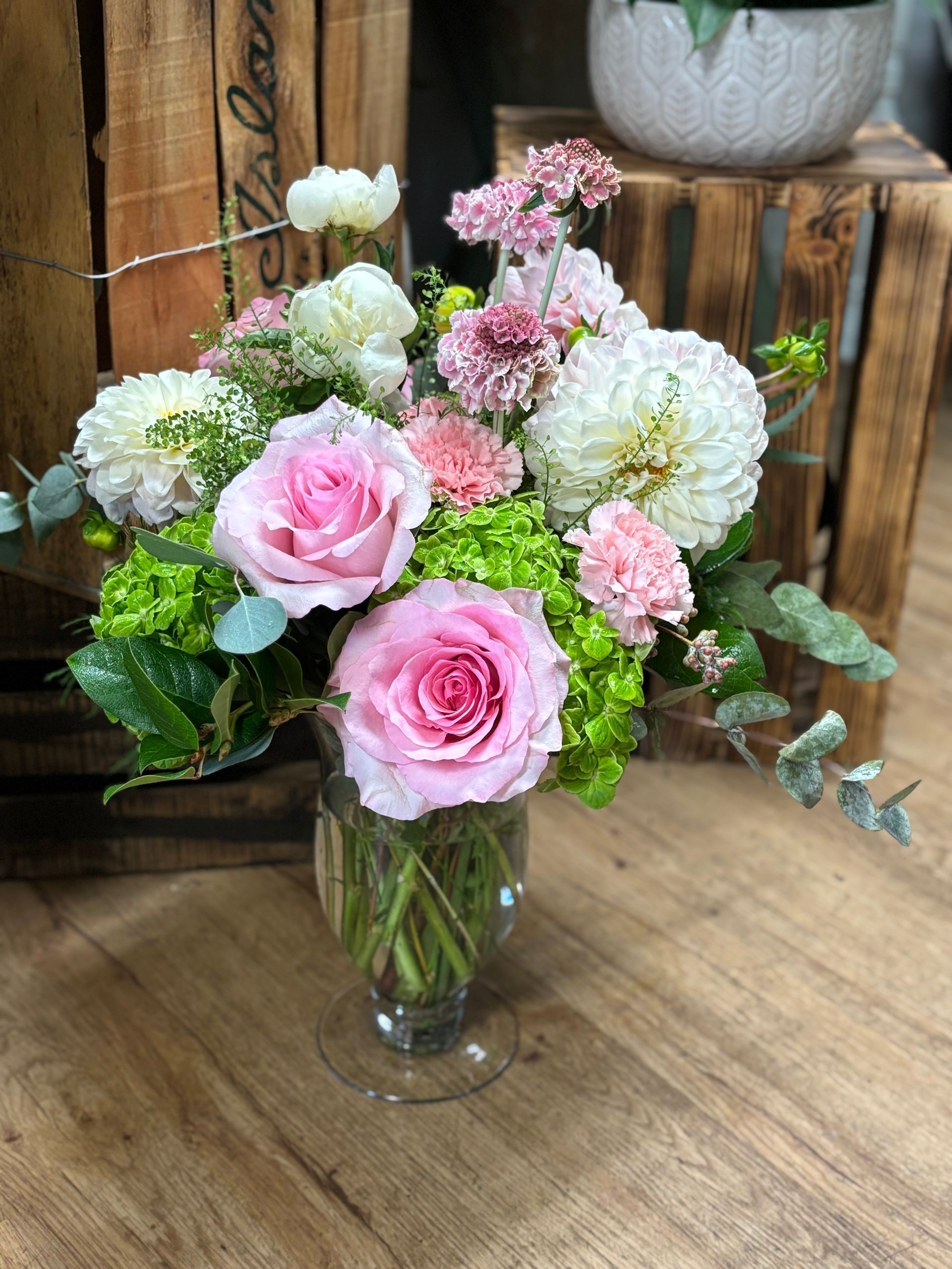 A glass vase containing a floral arrangement of pink roses, white dahlias, green hydrangeas, and eucalyptus on wood.