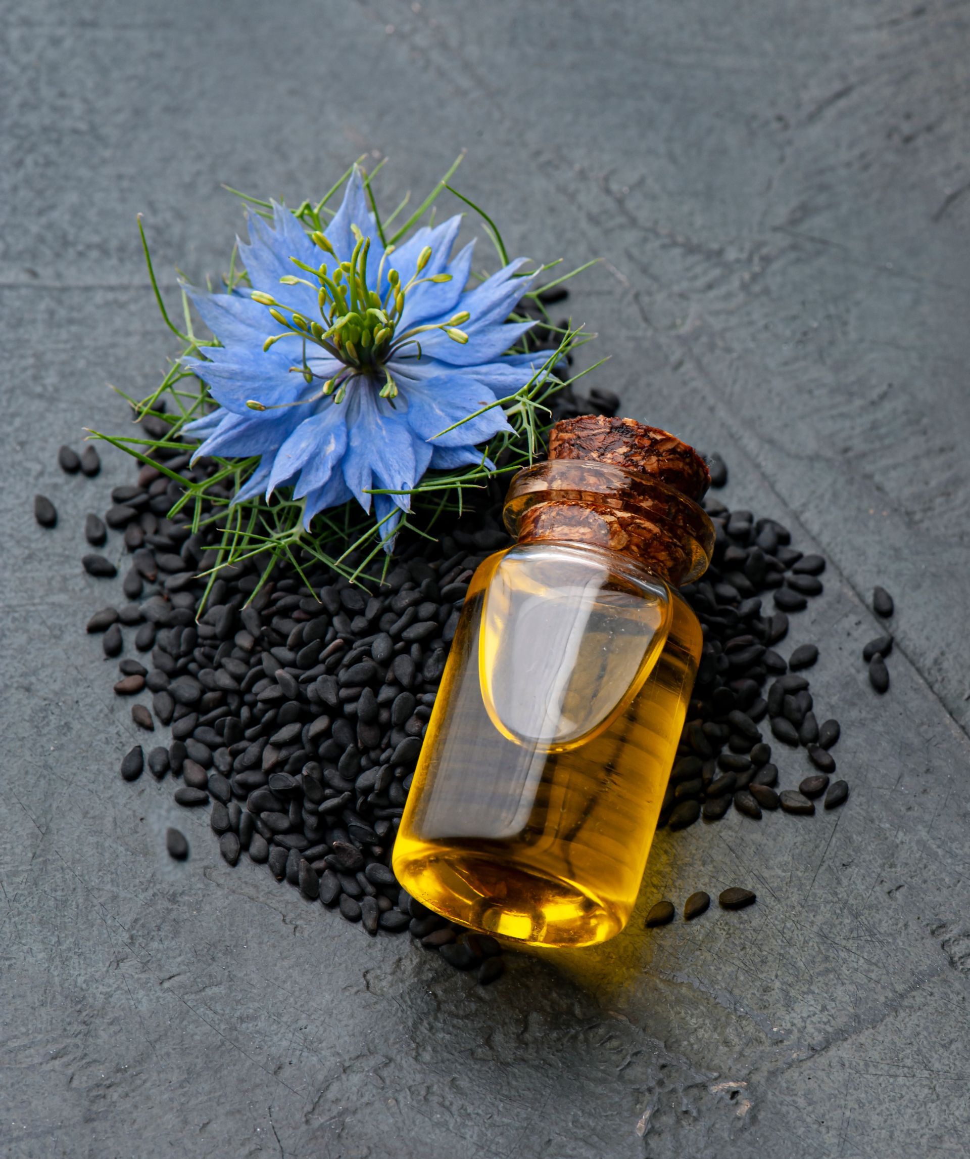 Une bouteille d'huile de nigelle à côté d'une fleur bleue et de graines noires.