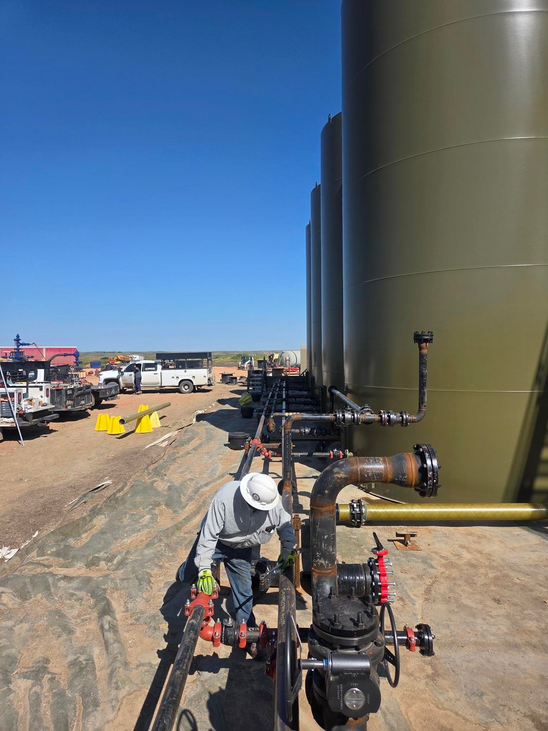 Worker in hard hat inspecting industrial pipes beside a large green storage tank at a construction site
