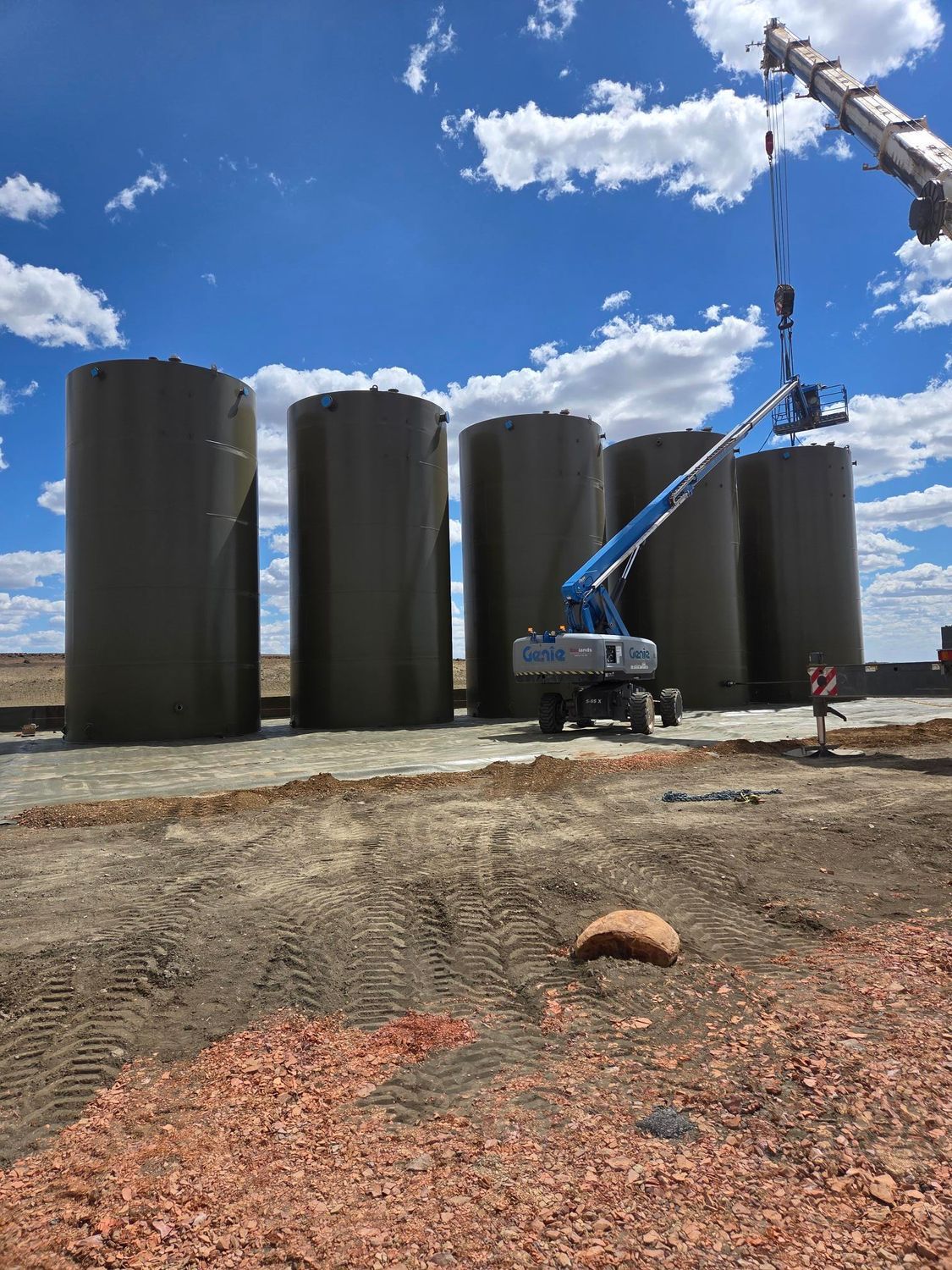 Blue crane beside four tall black storage tanks on a gravel site under a partly cloudy sky