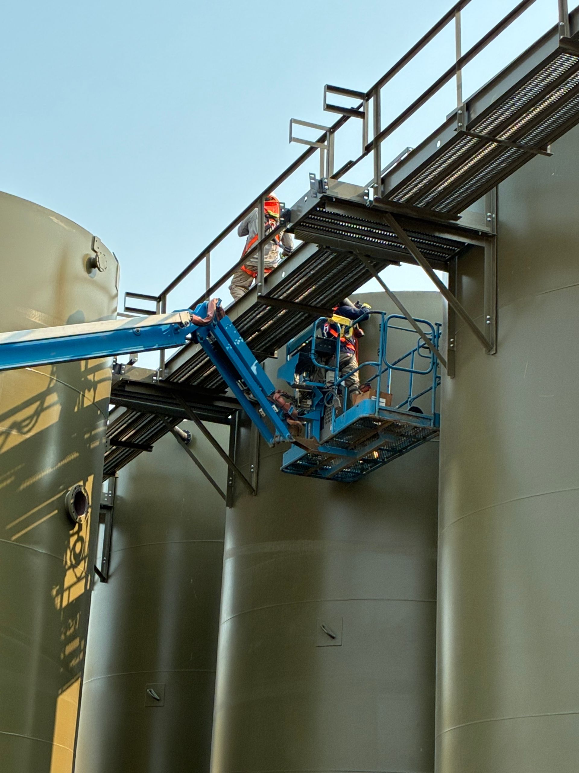 Worker in orange safety gear on a blue boom lift beside large industrial tanks and stairs