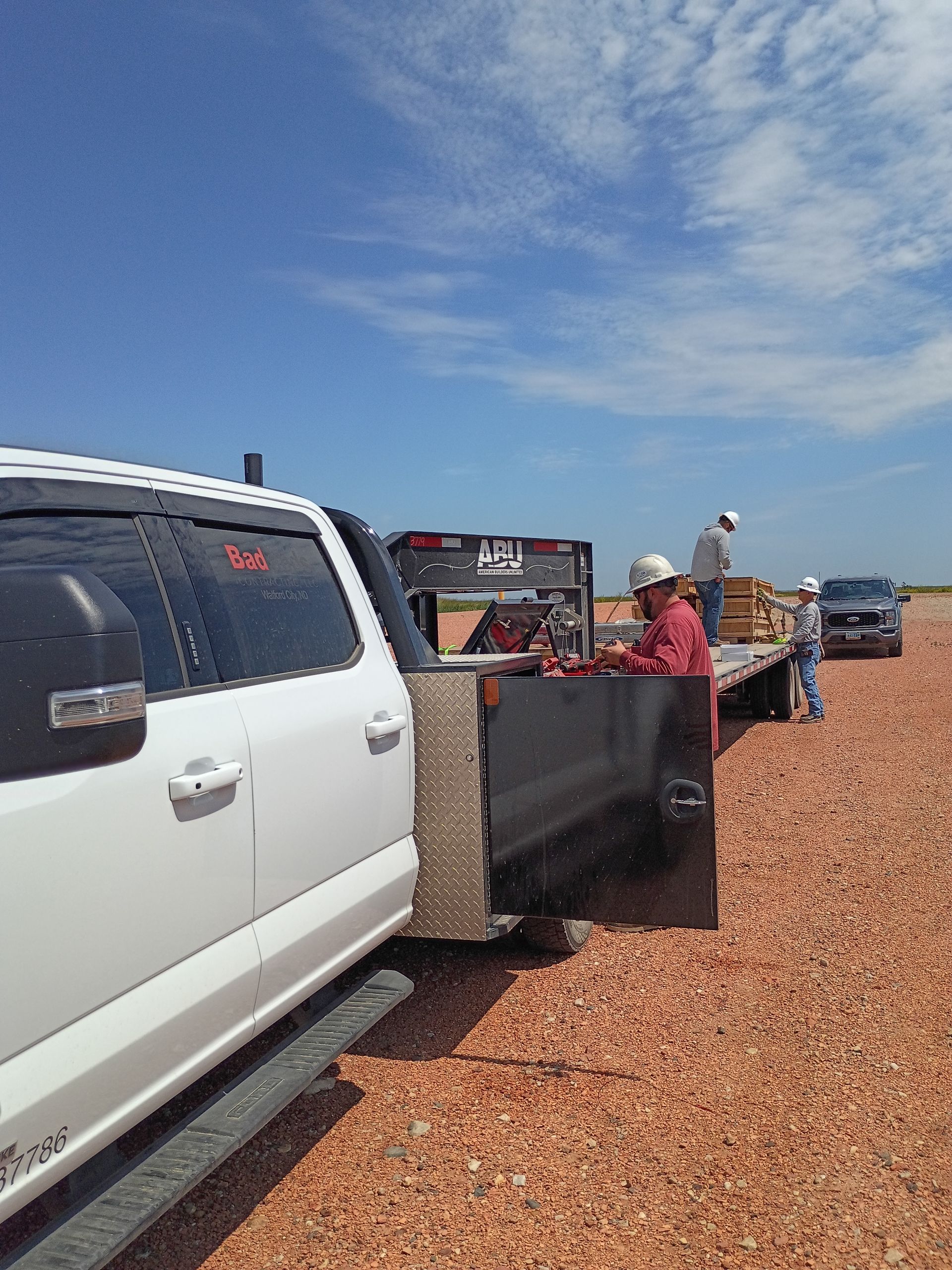 White SUV and black pickup parked on a gravel lot under a blue sky, with people near a trailer and tents.