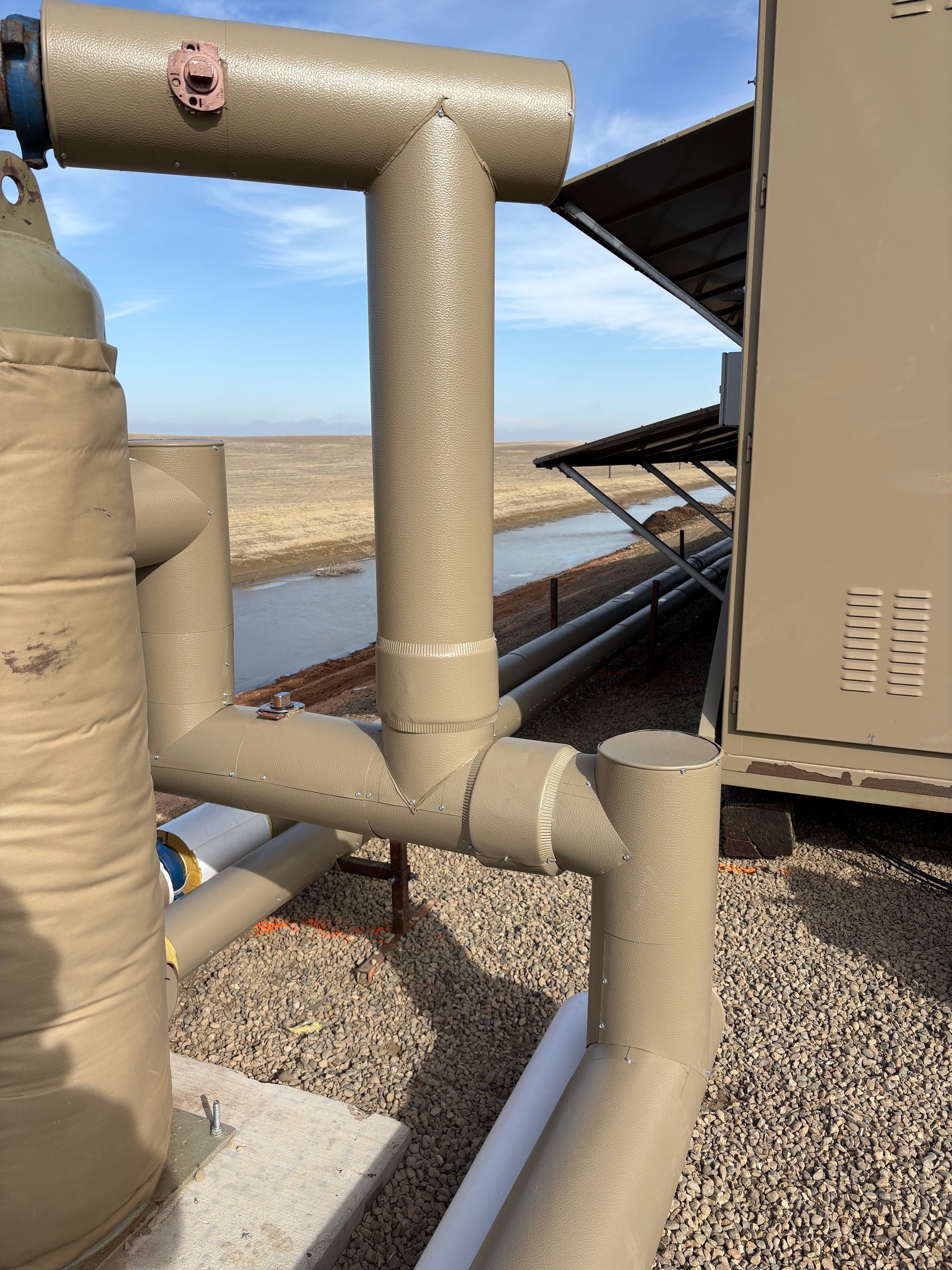 Beige industrial pipes and valves beside a gravel area, with a sloped metal structure in the background.
