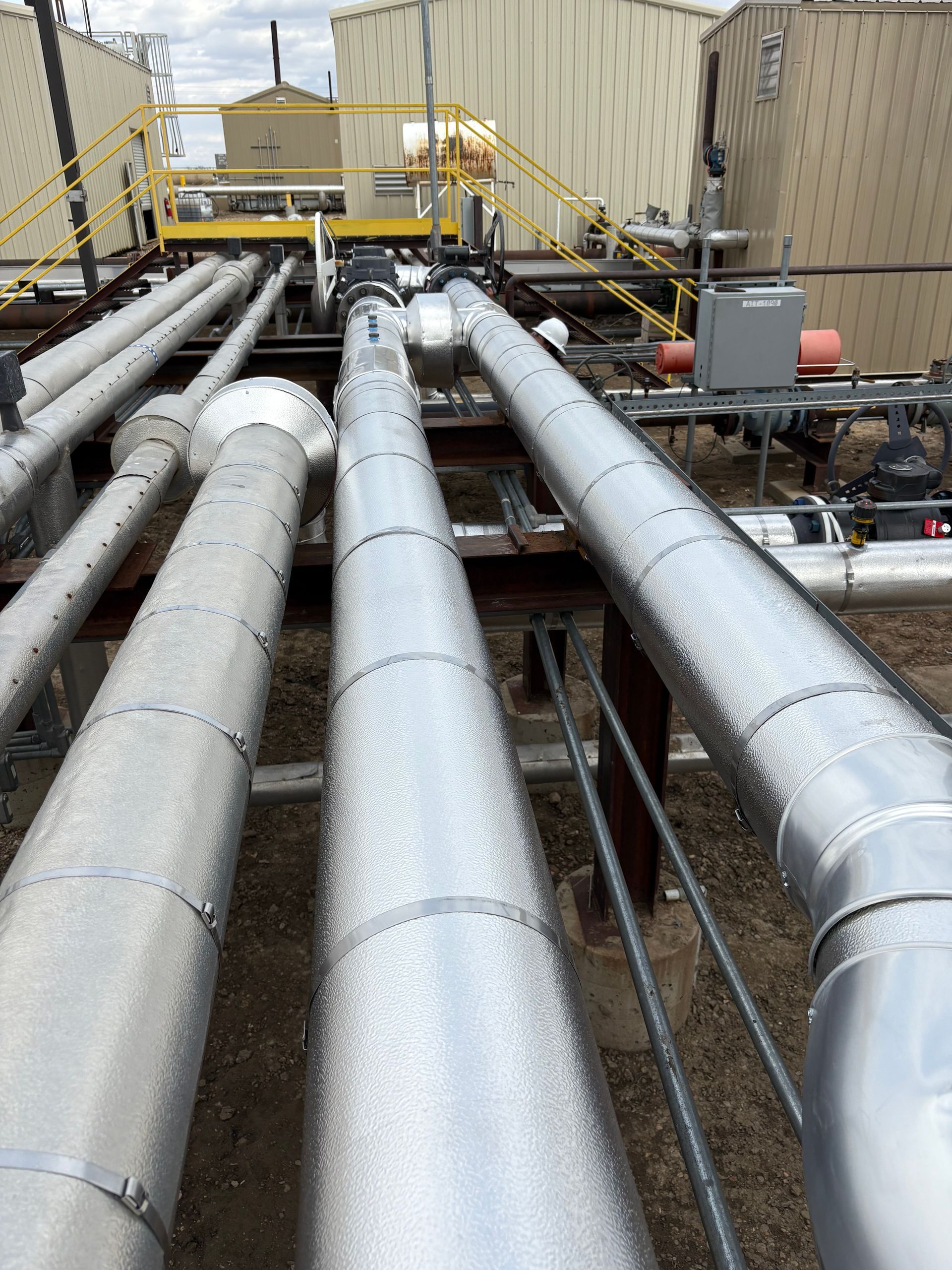 Industrial rooftop with large silver pipes and ducts leading toward beige utility buildings