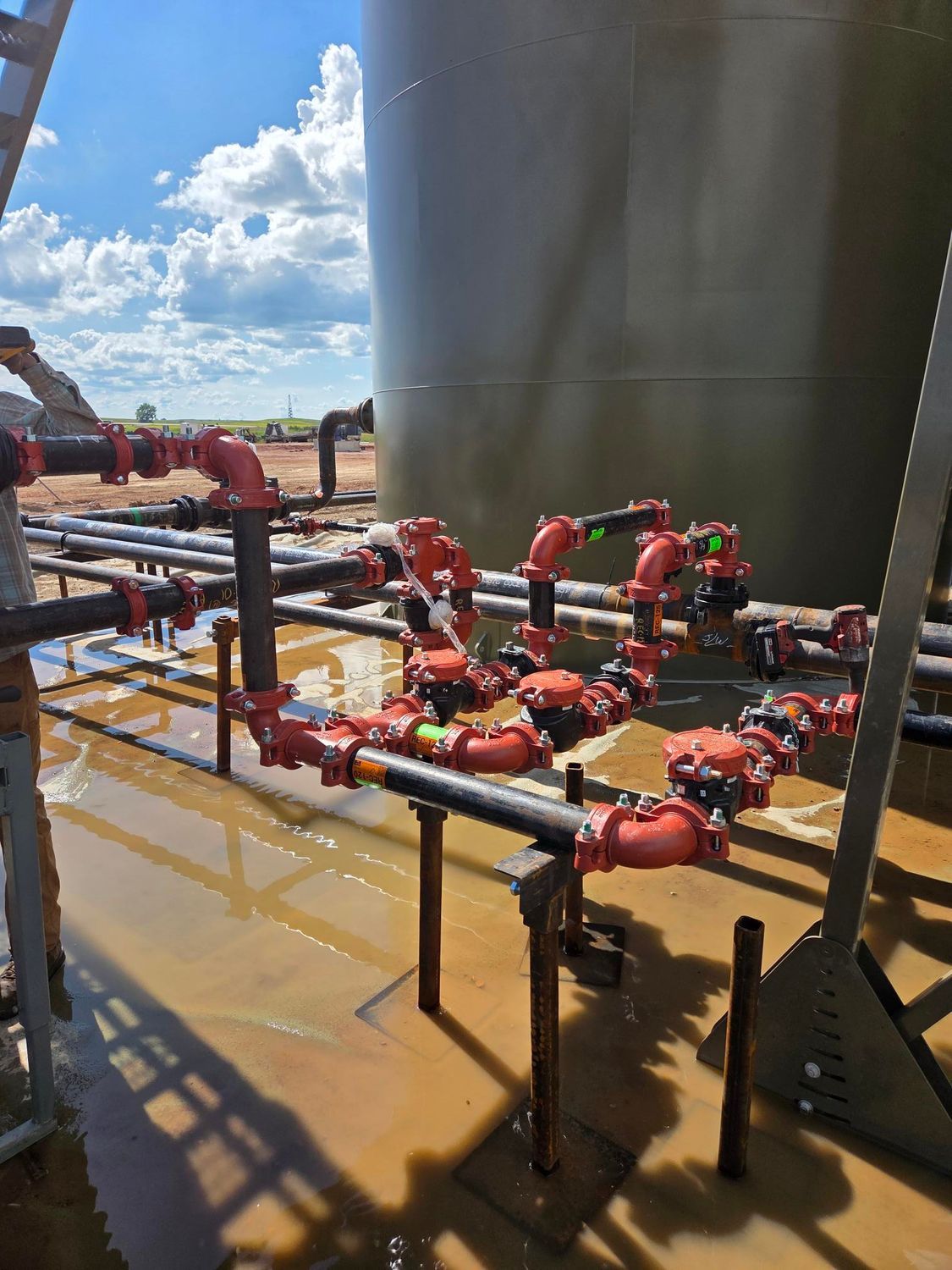 Industrial piping and red valves at a water facility beside a large tank, with wet ground and blue sky